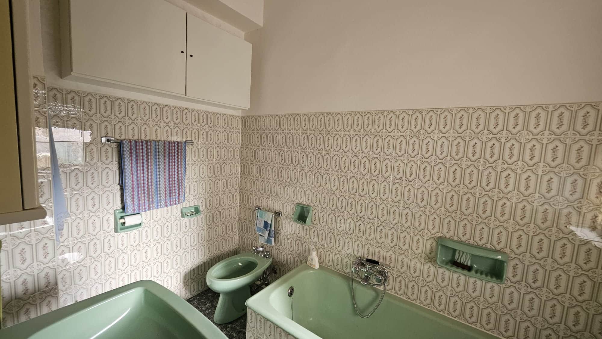 Vecchio bagno con piastrelle beige con motivi floreali, vasca e bidet verde acqua, armadietto bianco e accessori vintage, atmosfera nostalgica e caratteristica, ideale per amanti dell'arredamento d'epoca.