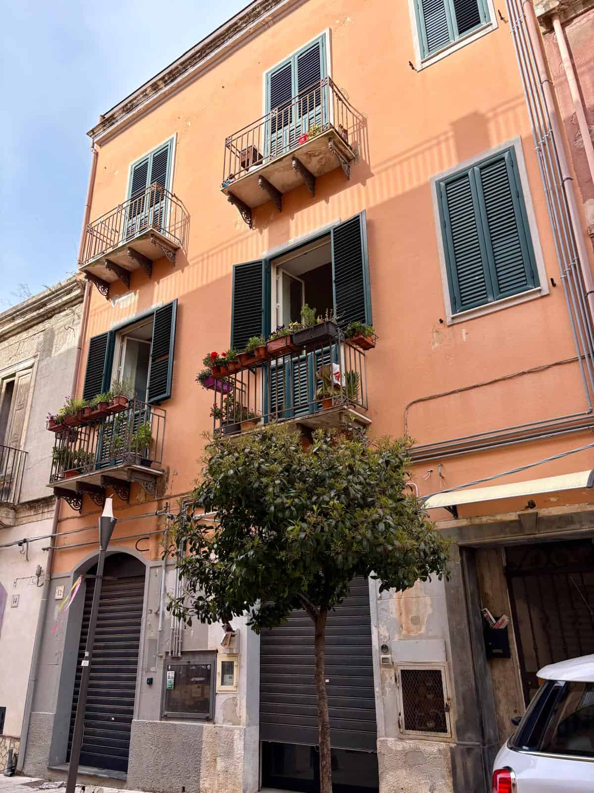 Balconi di un edificio residenziale con piante e fiori, facciata in colore pesca, finestre con persiane verdi, atmosfera urbana, in stile mediterraneo, immobile caratteristico, NextCasa.