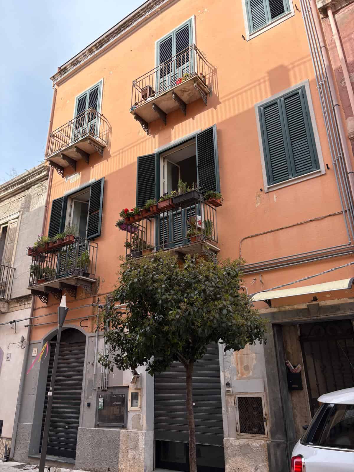 Fioreggiata abitazione con balconi in stile classico, facciata color pesca e finestre con tende a persiana blu, in zona urbana con auto parcheggiata e piante decorative sul balcone.