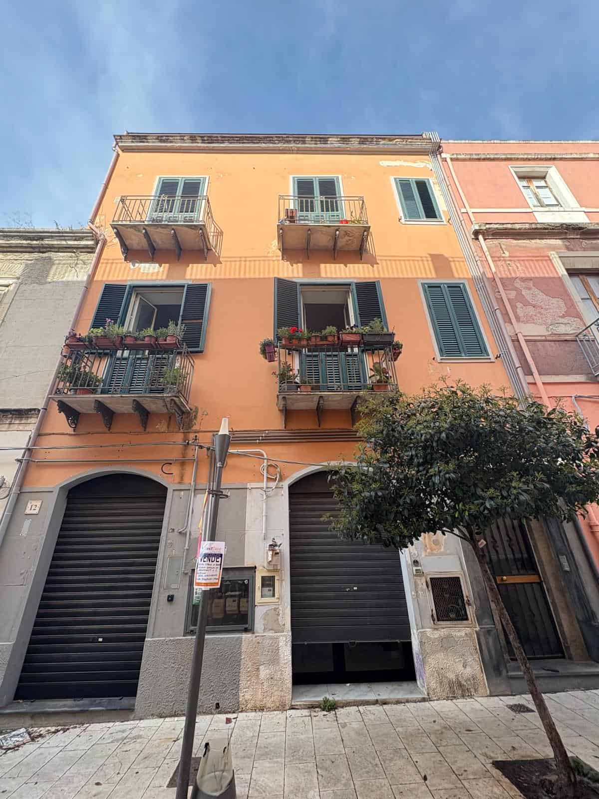 Immagine di un edificio residenziale di tre piani con facciata in tonalità arancione e finestre con persiane verdi, balconi con piante e fiori, situato in zona urbana con strada pedonale e albero in primo piano.