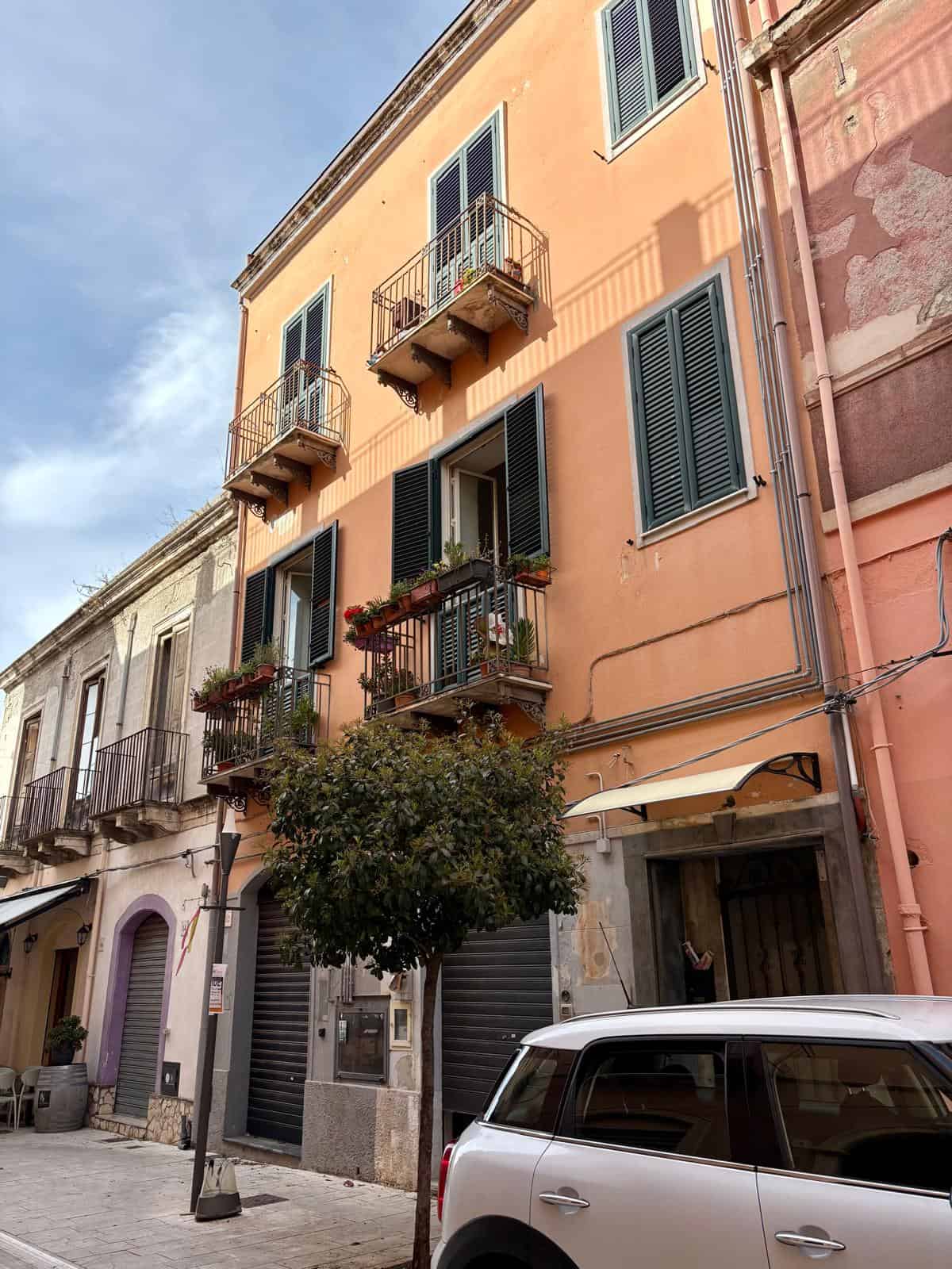 Balconi eleganti su palazzina storica nel centro di Napoli, con fioriere e finestre con persiane verdi, in una strada vivace con auto e negozi, rappresentando il fascino delle case italiane.