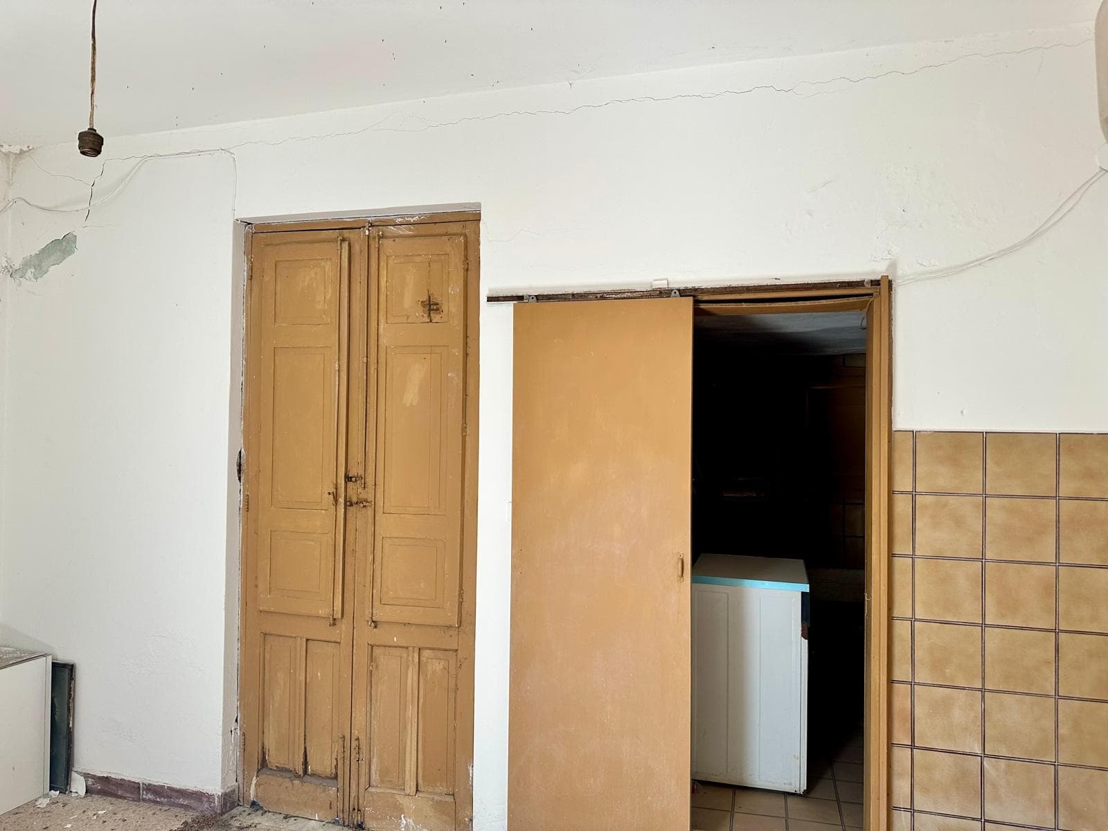 Vecchio armadio in legno con porte scorrevoli, ingresso alla cucina con porta apribile, pareti bianche, rivestimento di ceramica marrone chiaro, soffitto e pareti in cattive condizioni, spazio di ristrutturazione.