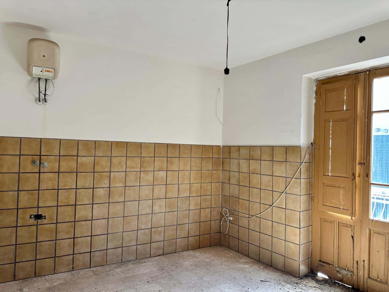 Parete cucina in fase di ristrutturazione con piastrelle beige, grande finestra in legno, presa e attacchi elettrici pronti per gli impianti, ambienti da personalizzare.