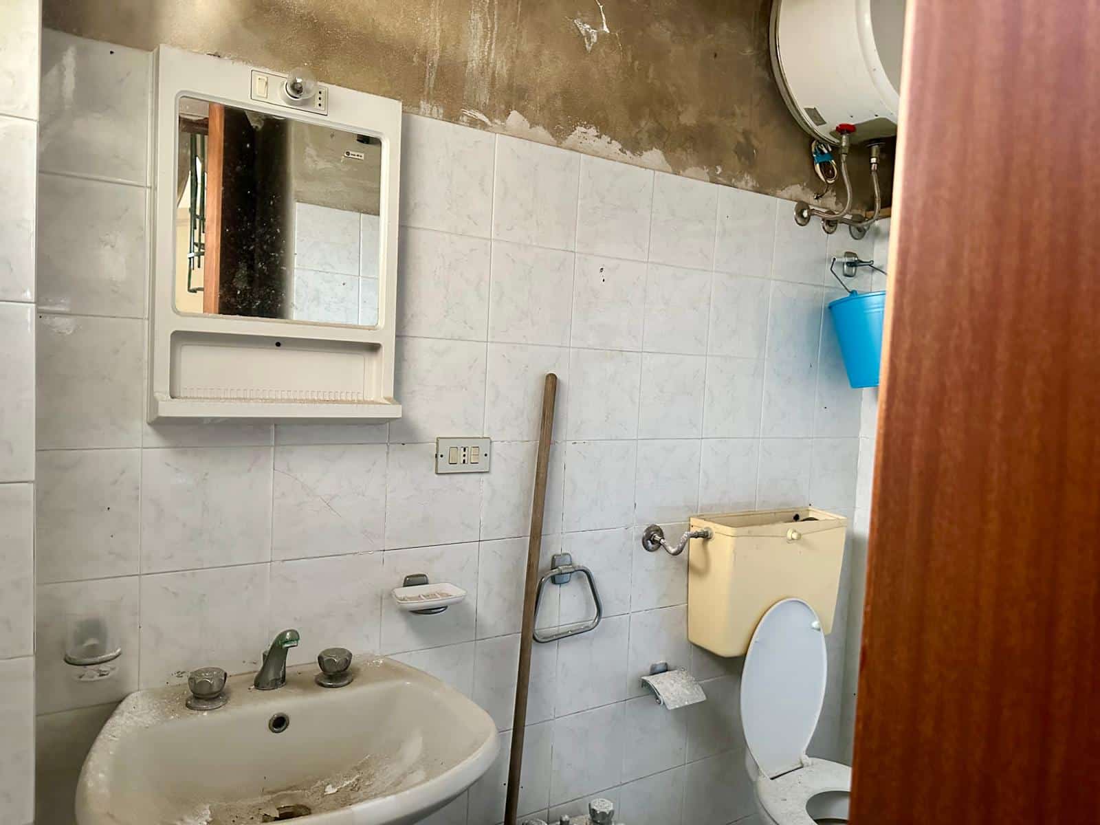 Lavabo e specchio in bagno modesto con pareti piastrellate e elementi di arredo di vecchia data, presenza di secchio blu e attrezzature di servizio, ambiente funzionale ma datato per ristrutturazione.