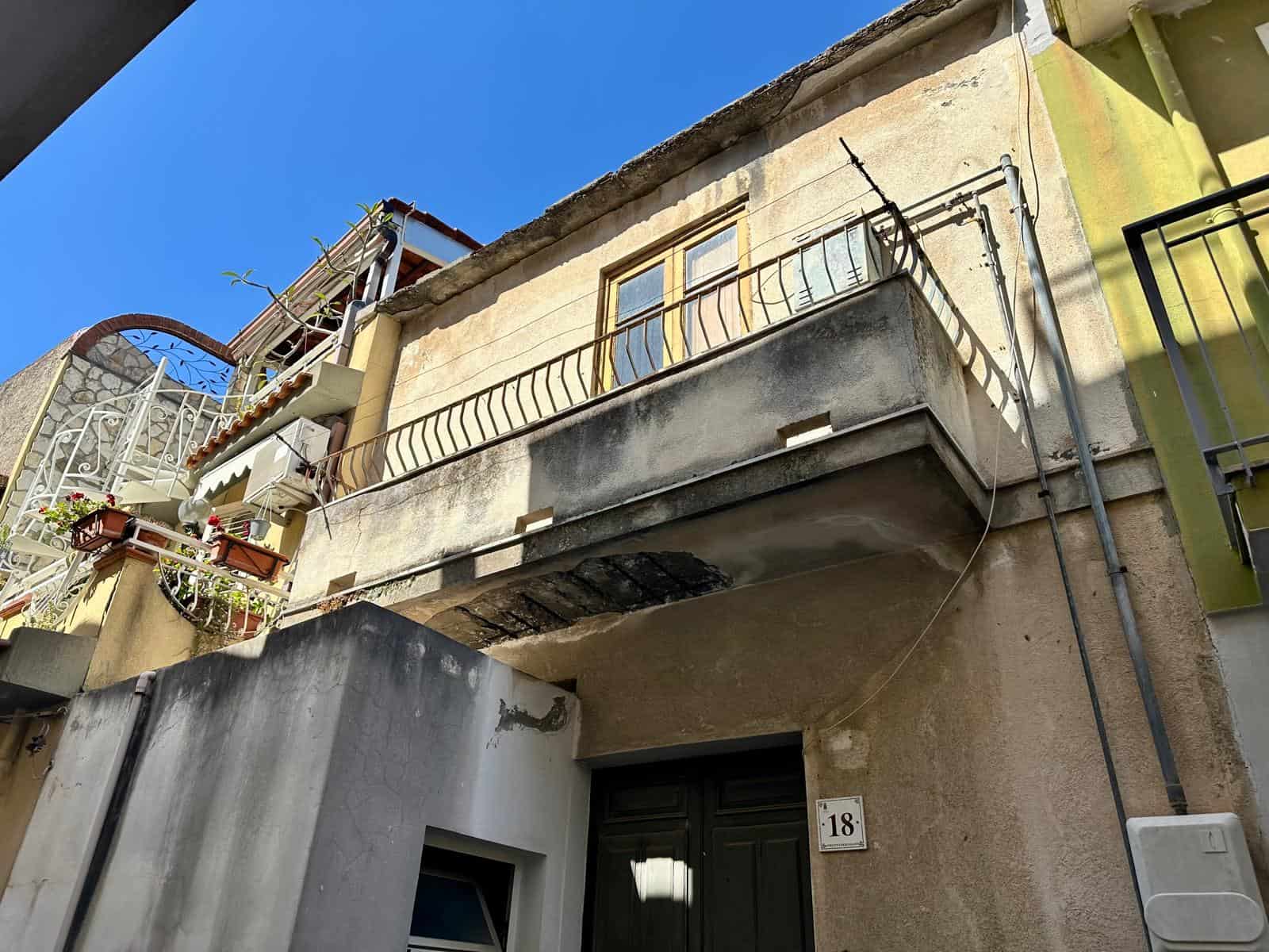 Balcone di un edificio storico con ringhiera in ferro battuto, pareti in pietra e soffitto in cemento. Facciata esterna con piante e condizioni dell'edificio in zona urbana, sotto un cielo limpido.