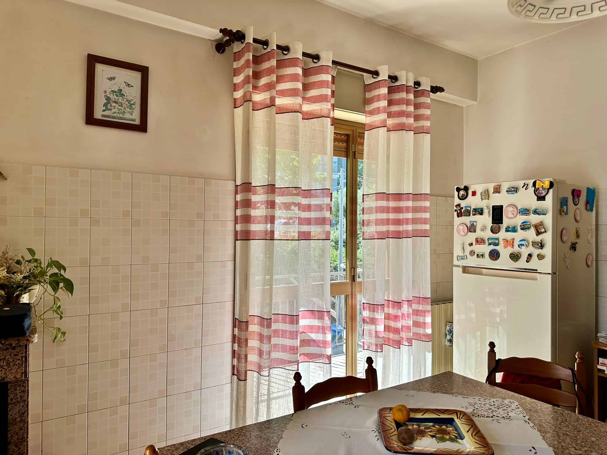 Vista cucina con tendaggi a righe, frigorifero decorato con calamite, ambiente accogliente e luminoso, elementi di arredo tradizionali, ideale per la vendita immobiliare.