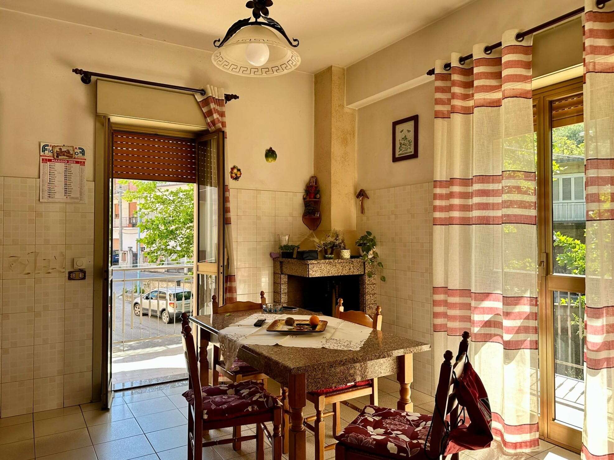Finestra con tende a righe rosse e bianche in cucina con vista su strada e automobili, ambiente caldo e accogliente con pareti beige, camino e arredamento in stile tradizionale.