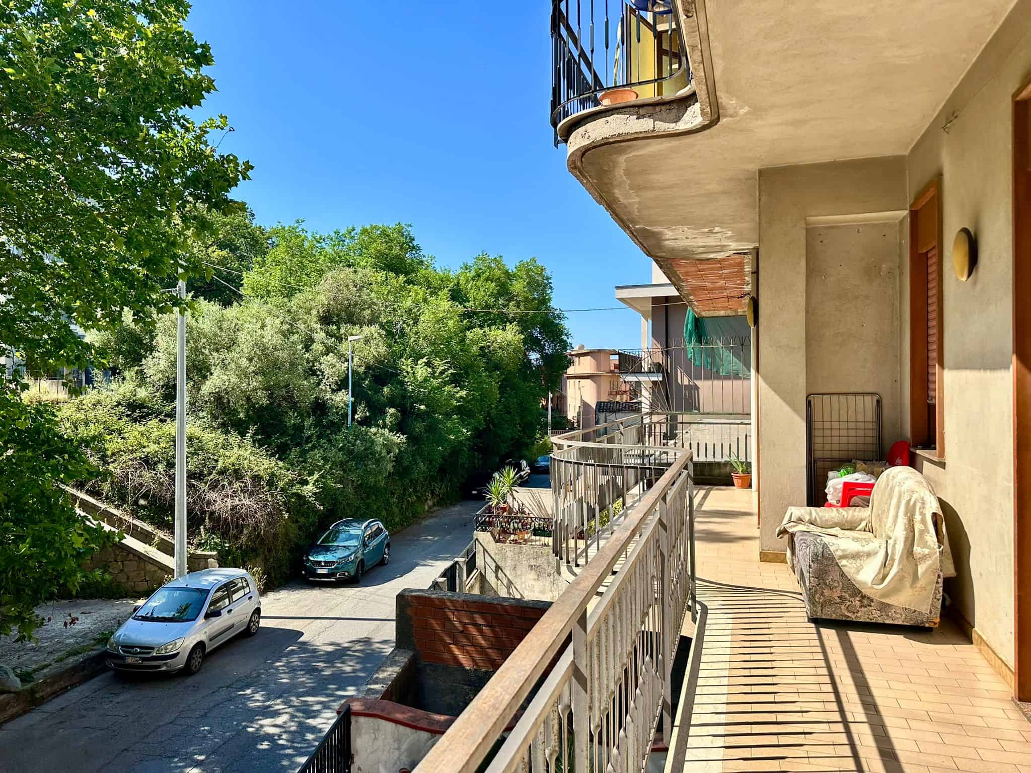 Balcone di un appartamento con vista su strada alberata e parcheggi, con mobili e piante, sotto un cielo azzurro.