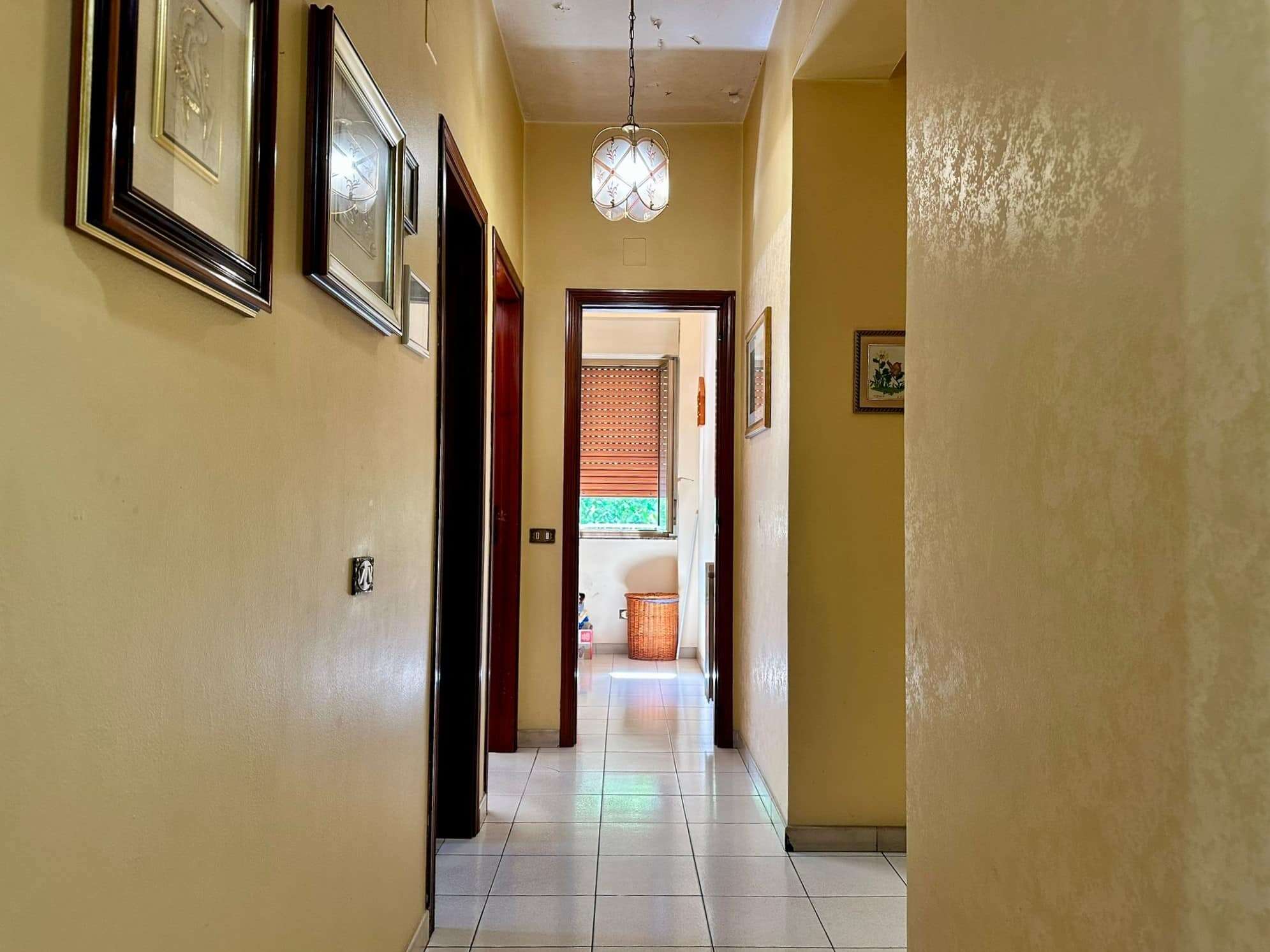 Spazioso corridoio in appartamento con pareti color crema e decorazioni di quadri, illuminato da lampadario vintage, vista verso una stanza con finestra, arredato in stile classico.
