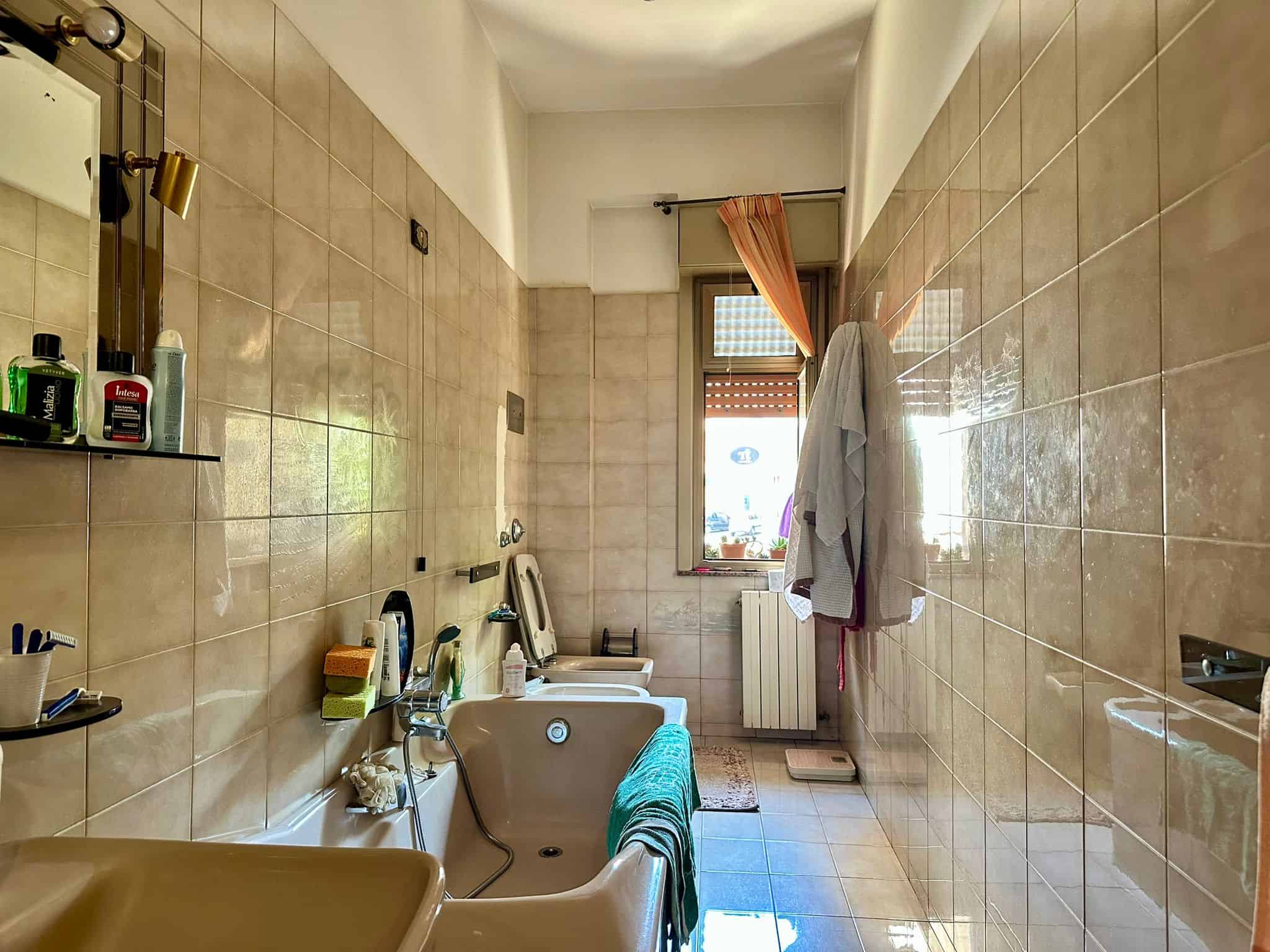 Bathroom con vasca e bidet, con pareti e pavimenti rivestiti in piastrelle beige, finestra con tende arancioni e asciugamani appesi, ambiente spazioso e luminoso.