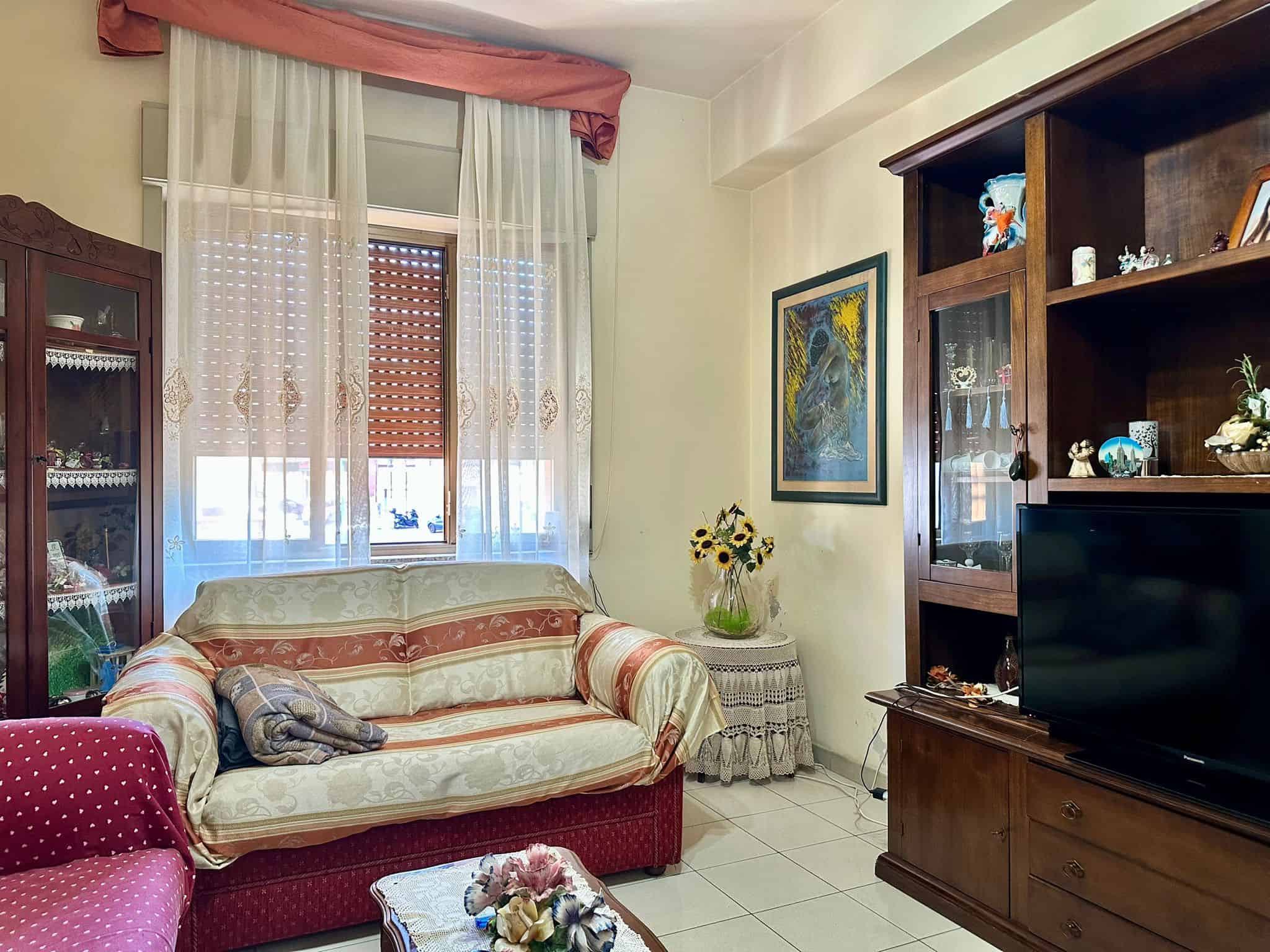 Soggiorno arredato con divani e mobili in legno, luci naturali da finestra decorata con tende leggere e dettagli floreali, in stile classico e accogliente.