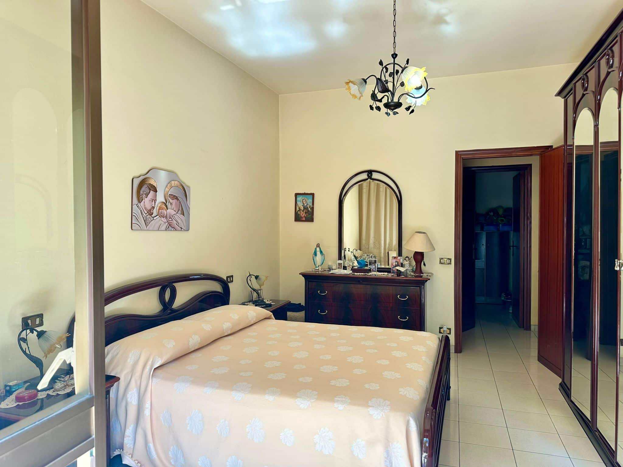 Letto matrimoniale in una camera da letto con mobili in legno scuro, quadri e specchio decorativo, lampade da comodino e armadio, ambiente luminoso e accogliente, stile classico.