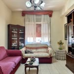 Divano rosso con poltrona in soggiorno, mobili di legno, tende leggere e decorazioni floreali, atmosfera accogliente e classica, ideale per living room confortevole e elegante.