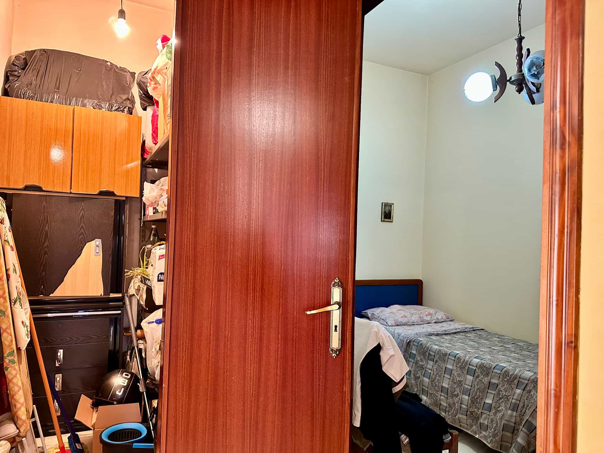 Armadio in legno aperto che rivela oggetti, vestiti e scatole in un ambiente domestico. Spazio disordinato con vari oggetti e arredi, vista sulla camera da letto con letto double e pareti chiare.