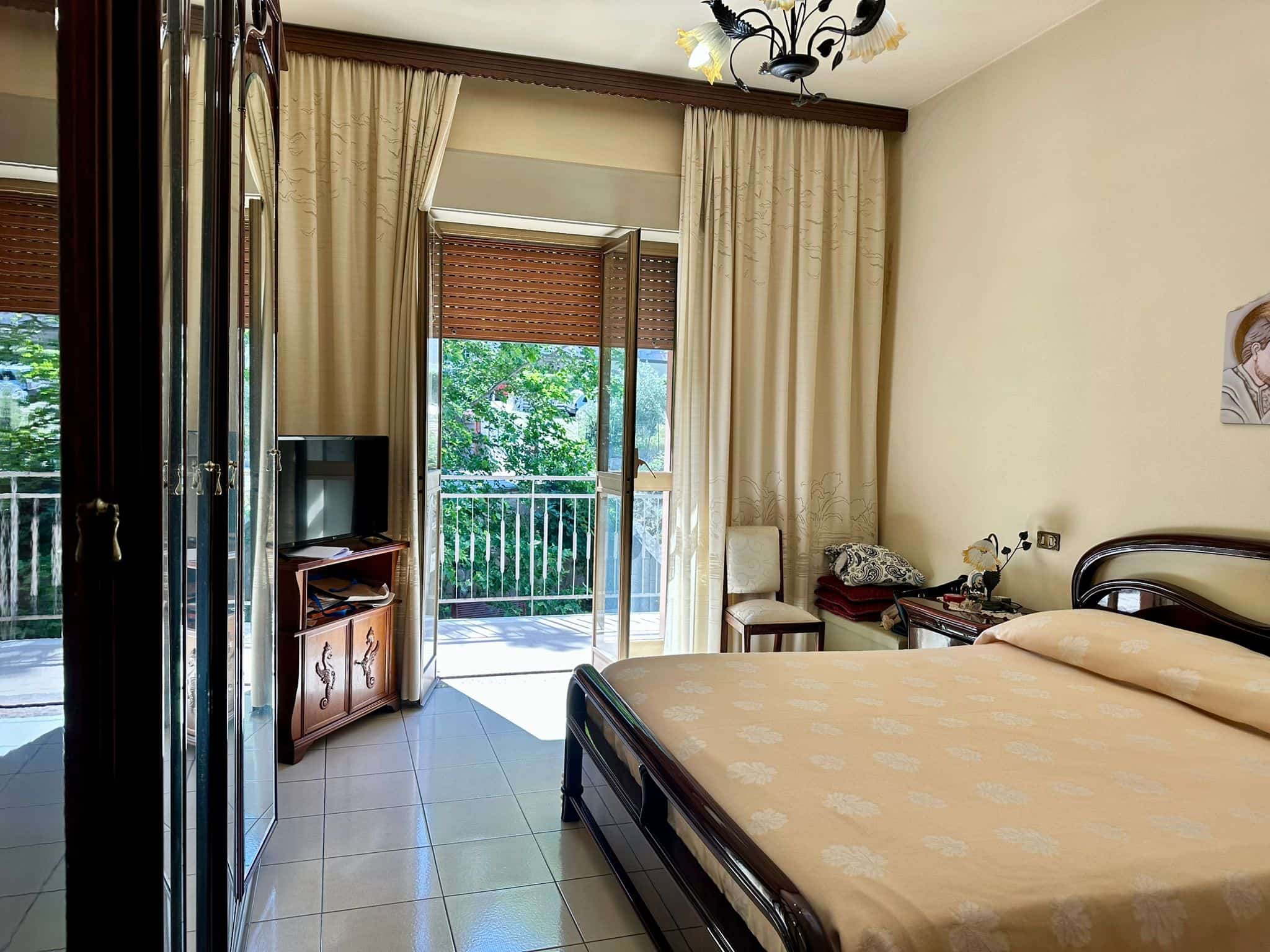 Spazioso appartamento con camera da letto matrimoniale che si affaccia su un balcone con vista sul verde, arredi eleganti, tende pesanti, e zona soggiorno luminosa.