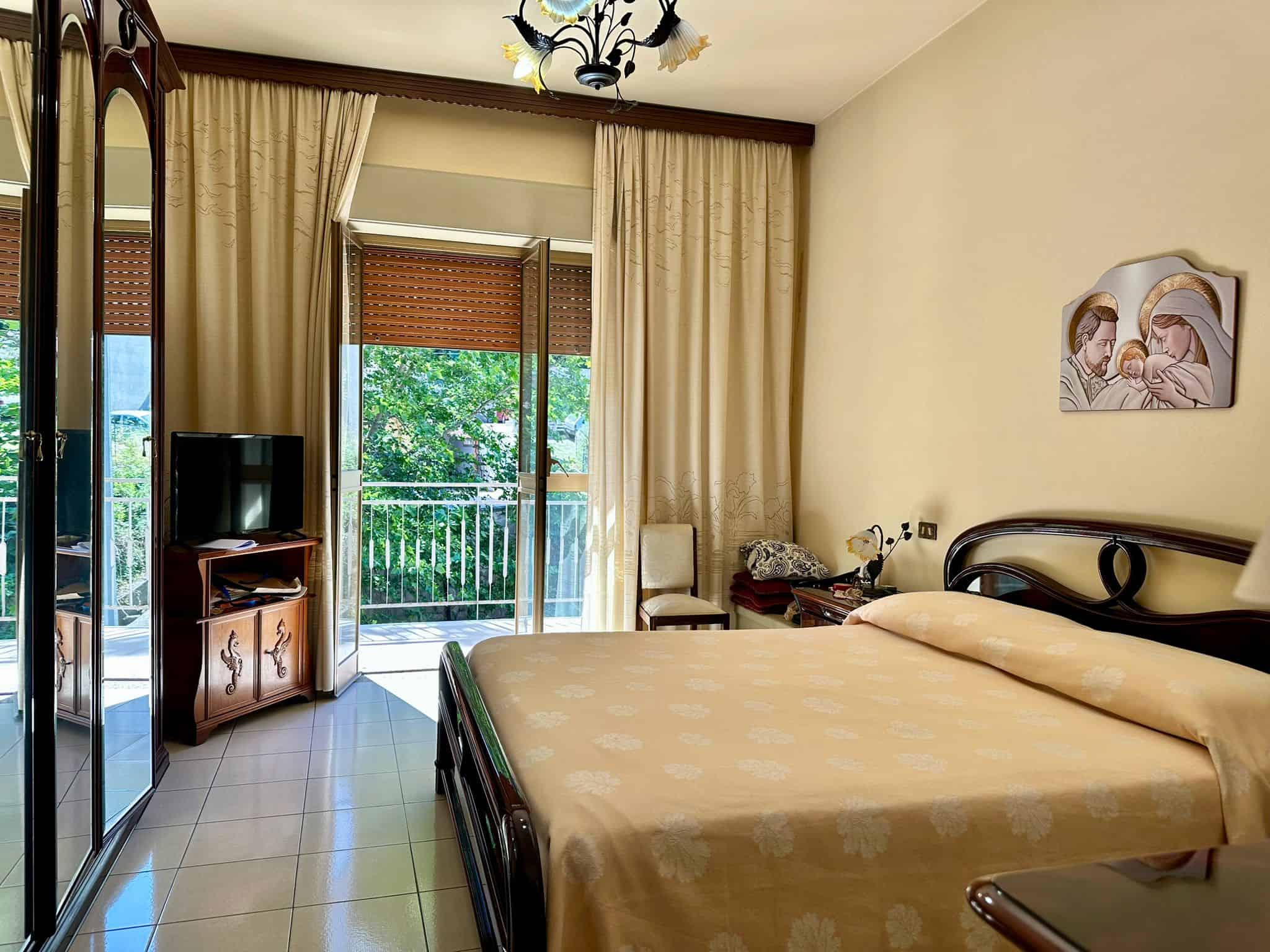 Splendida camera da letto matrimoniale con accesso al balcone, arredamento classico in legno, tendaggi eleganti e TV, ideale per affitti turistici di qualità a Giardini Naxos.