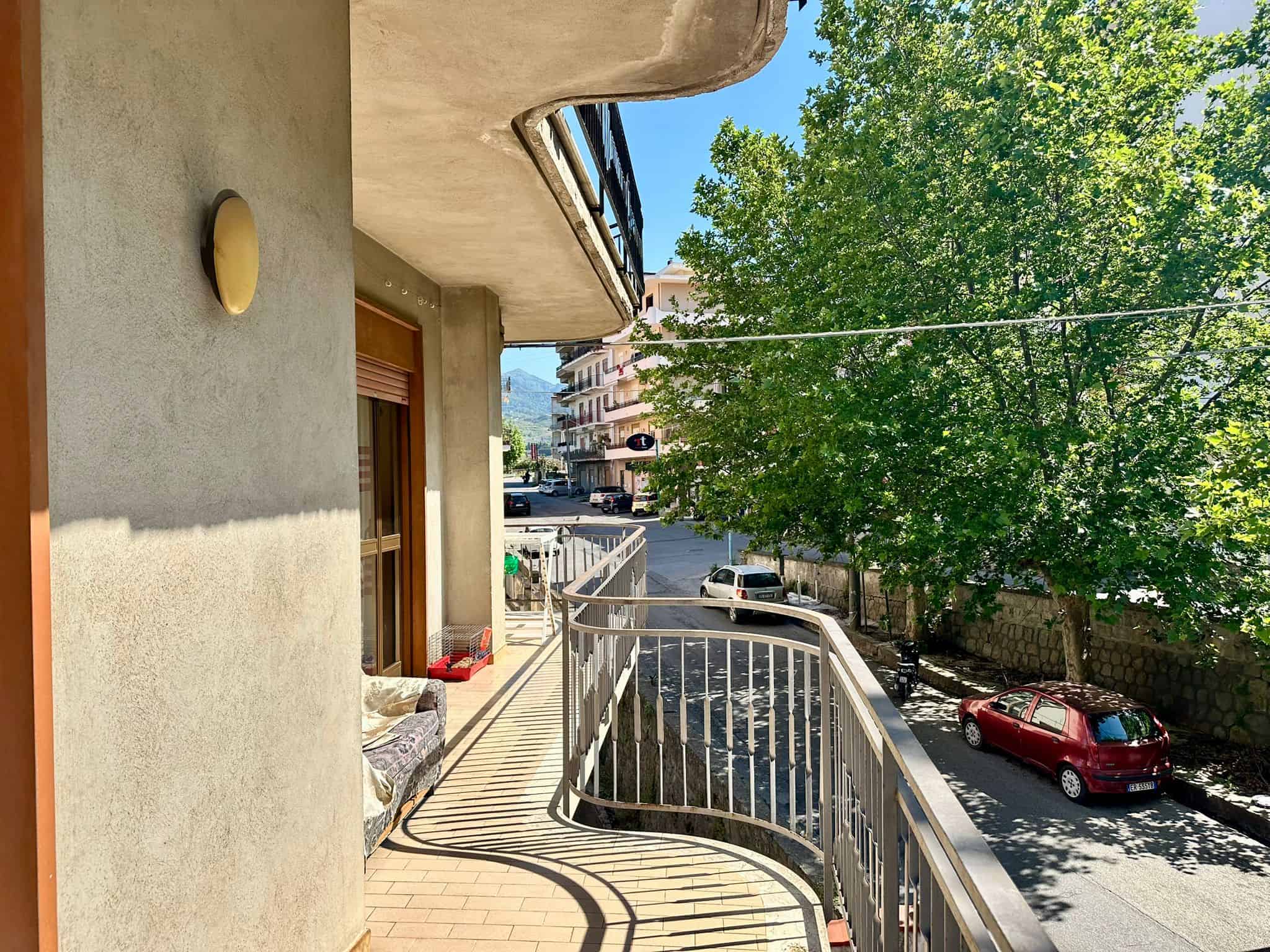 Vista balcone con ringhiera, strada con auto parcheggiate, alberi verdi, edifici residenziali e montagne sullo sfondo, illuminata dalla luce solare, atmosfera tranquilla in centro città.