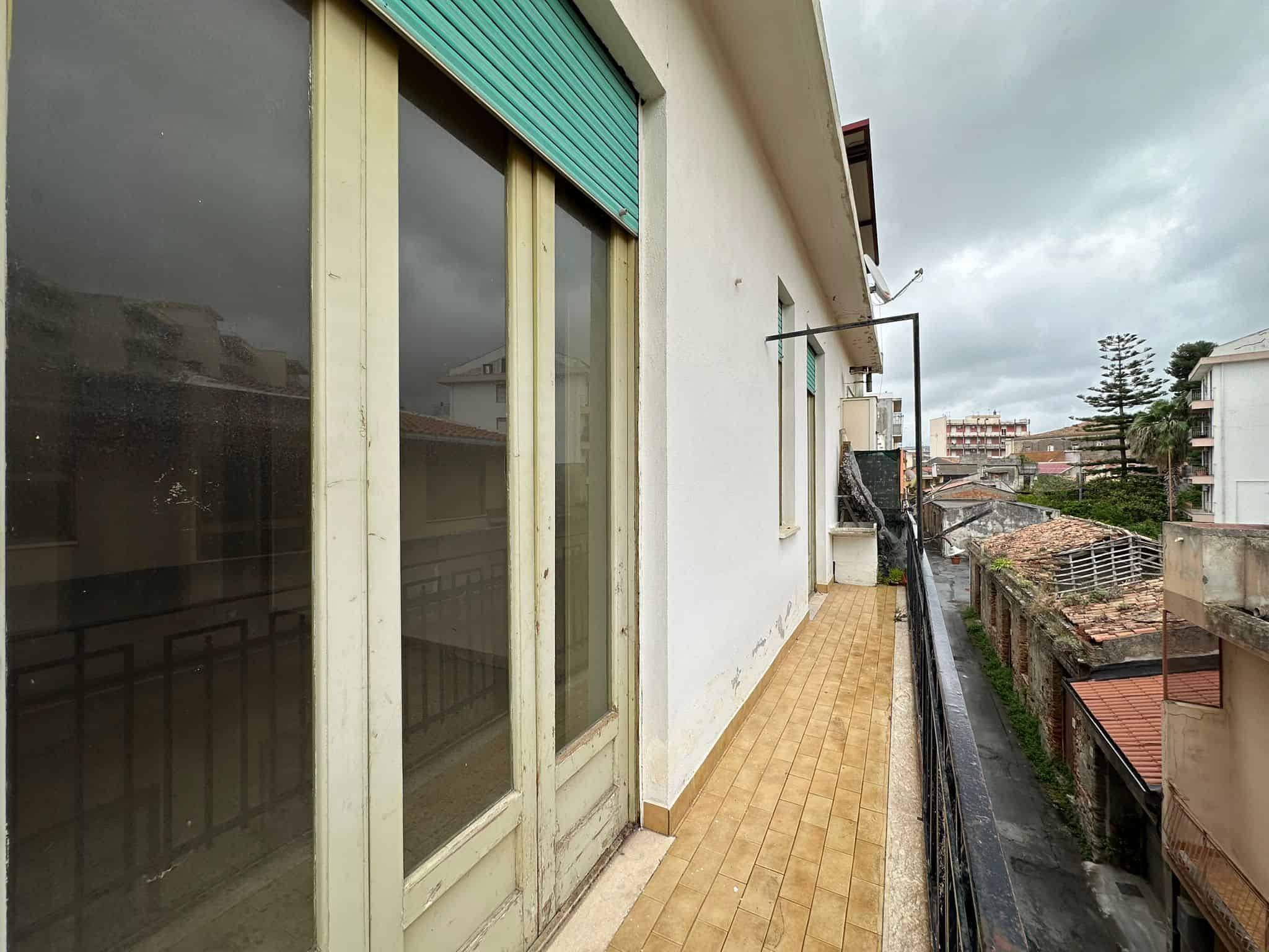 Balcone di un appartamento in città, vista urbana con case e alberi, pavimentazione in piastrelle beige, pareti bianche e finestre con tapparelle in plastica verde, cielo nuvoloso.