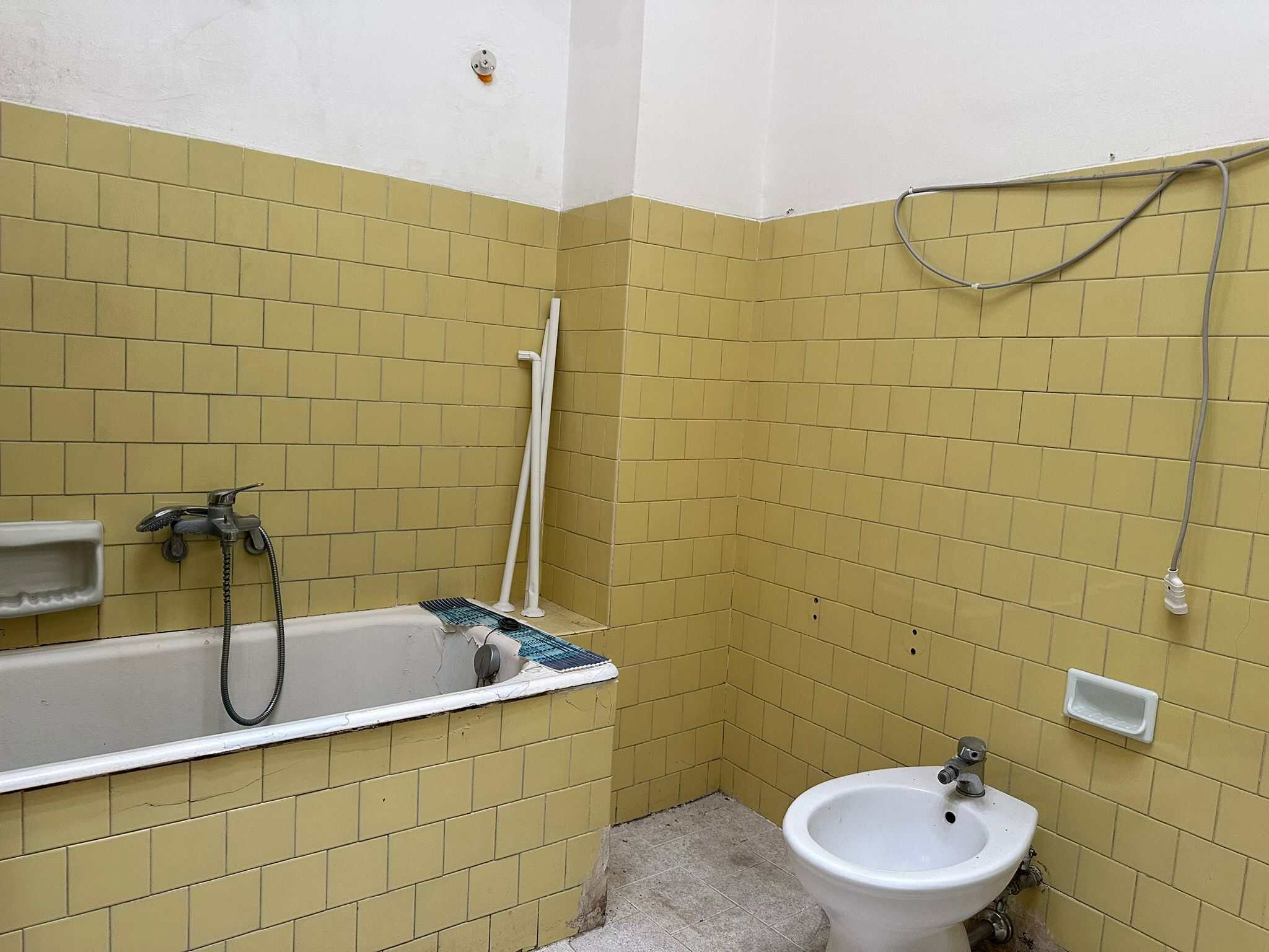 Vecchia stanza bagno con piastrelle gialle, vasca, bidet e elementi di arredo abbandonati, in fase di ristrutturazione o manutenzione, ambiente funzionale e semplice.