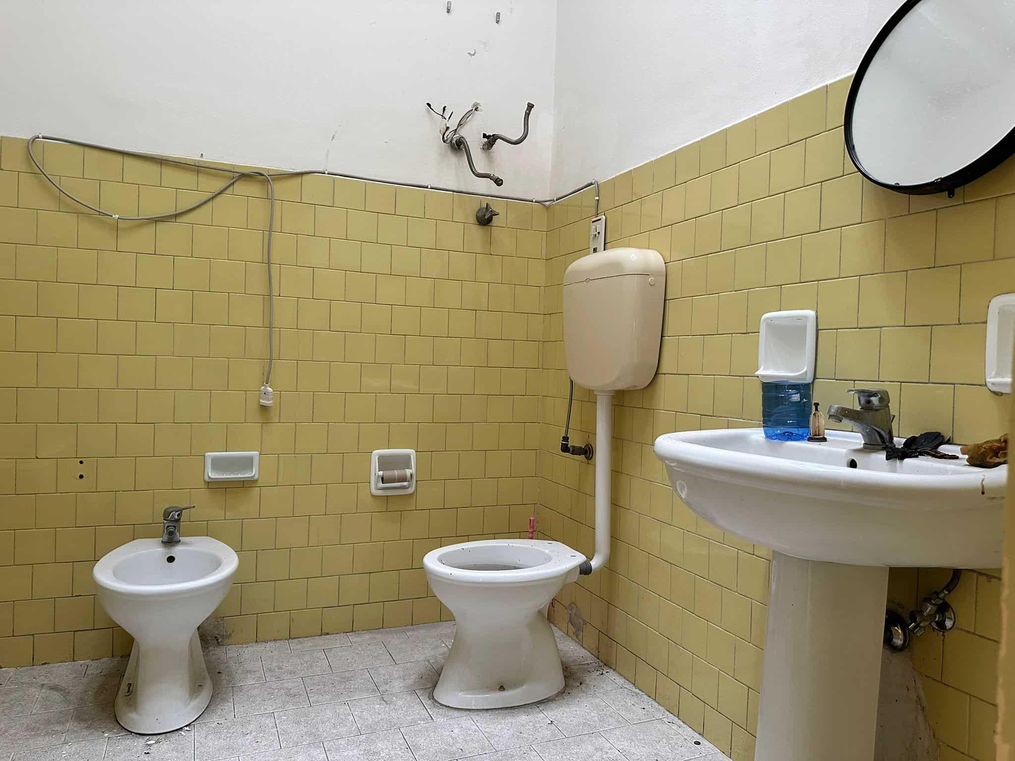 Bidet e WC in bagno con piastrelle gialle, lavabo con specchiera rotonda, ambiente di servizio in cattivo stato con cavi scoperti e mancanza di finiture, ambiente trasandato e da ristrutturare, bagno con arredi semplici e senza finiture moderne, spazio di servizio con elementi di edilizia datati e disorganizzati, immagini di un bagno vecchio non ristrutturato con caratteristiche da migliorare, l'area mostra evidente bisogno di manutenzione e rinnovamento.