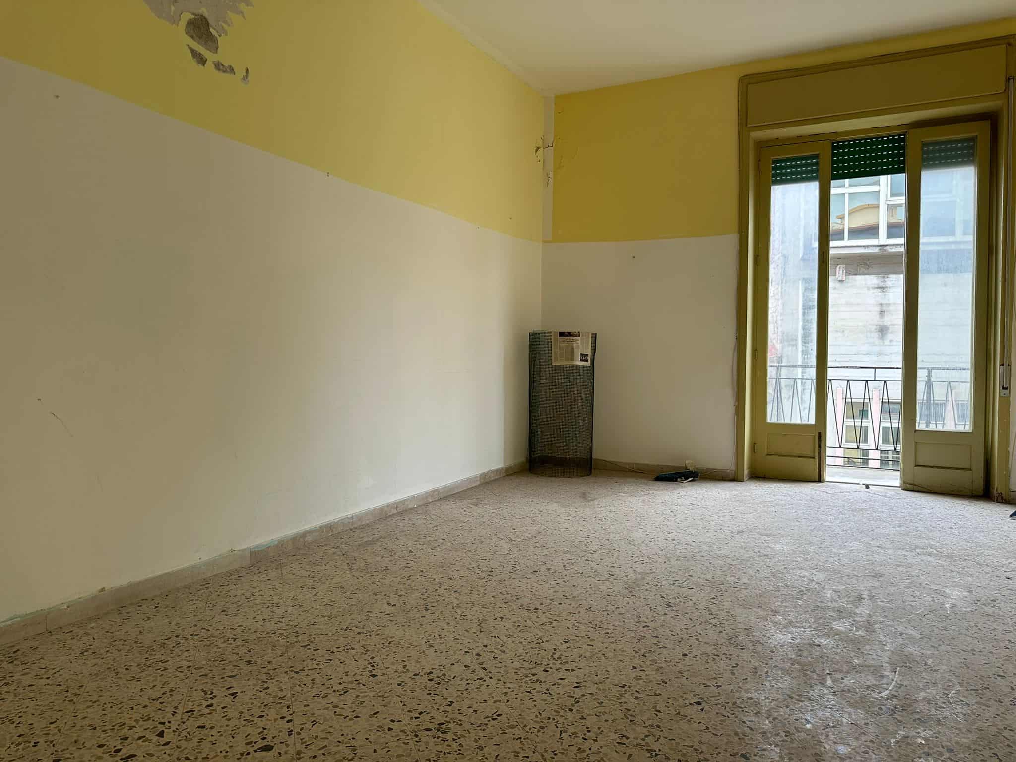 Soggiorno vuoto con pareti gialle e beige, grande finestra con portafinestra che conduce al balcone, pavimento in marmo, alloggio da ristrutturare, ambiente luminoso, potenziale di ristrutturazione, immobile in centro città, appartamento con vista, spazio abitativo da personalizzare.
