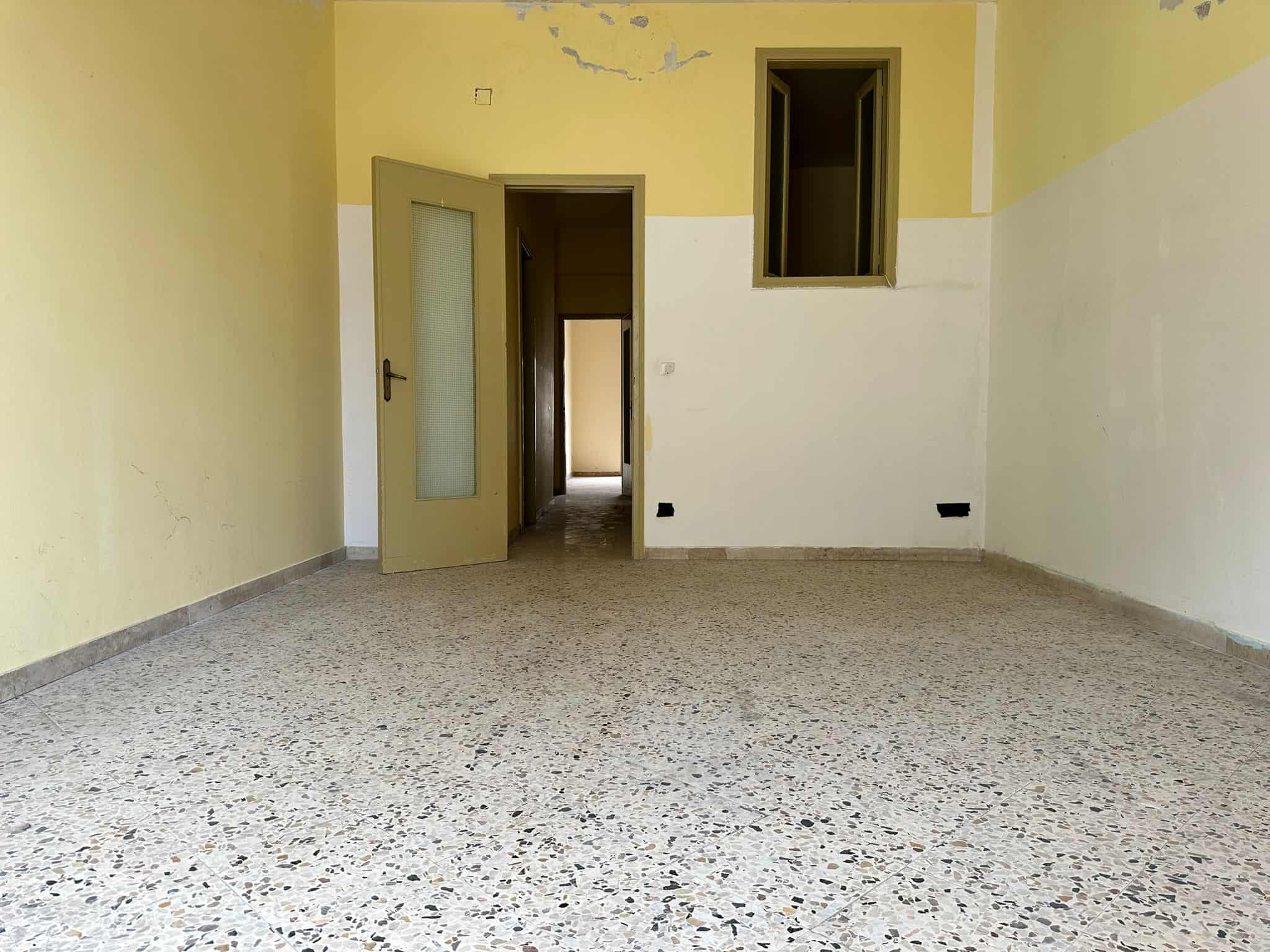 Pavimento in terrazzo in una casa in fase di ristrutturazione, pareti gialle e bianche, porta aperta che conduce ad altri ambienti, finestre piccole, aspetto giovane e luminoso.