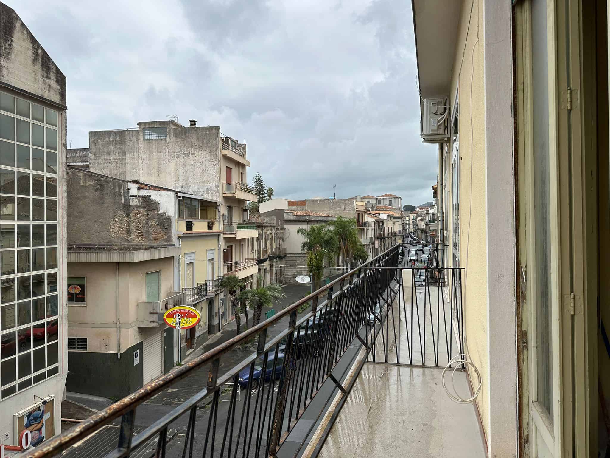 Balcone di un appartamento con vista su strada urbana, edifici storici e negozi, cielo nuvoloso, dettagli di condizionatori e ringhiera in ferro battuto, atmosfera metropolitana.