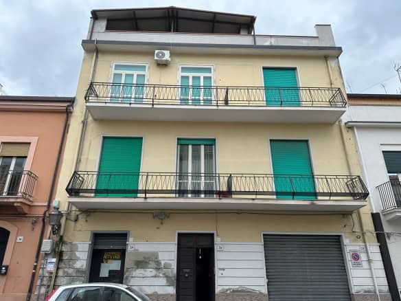 Balcone di un edificio residenziale con porte e finestre a vetri, persiane verdi, ringhiera in metallo e aria condizionata, situato in un'area urbana con altri edifici vicini.