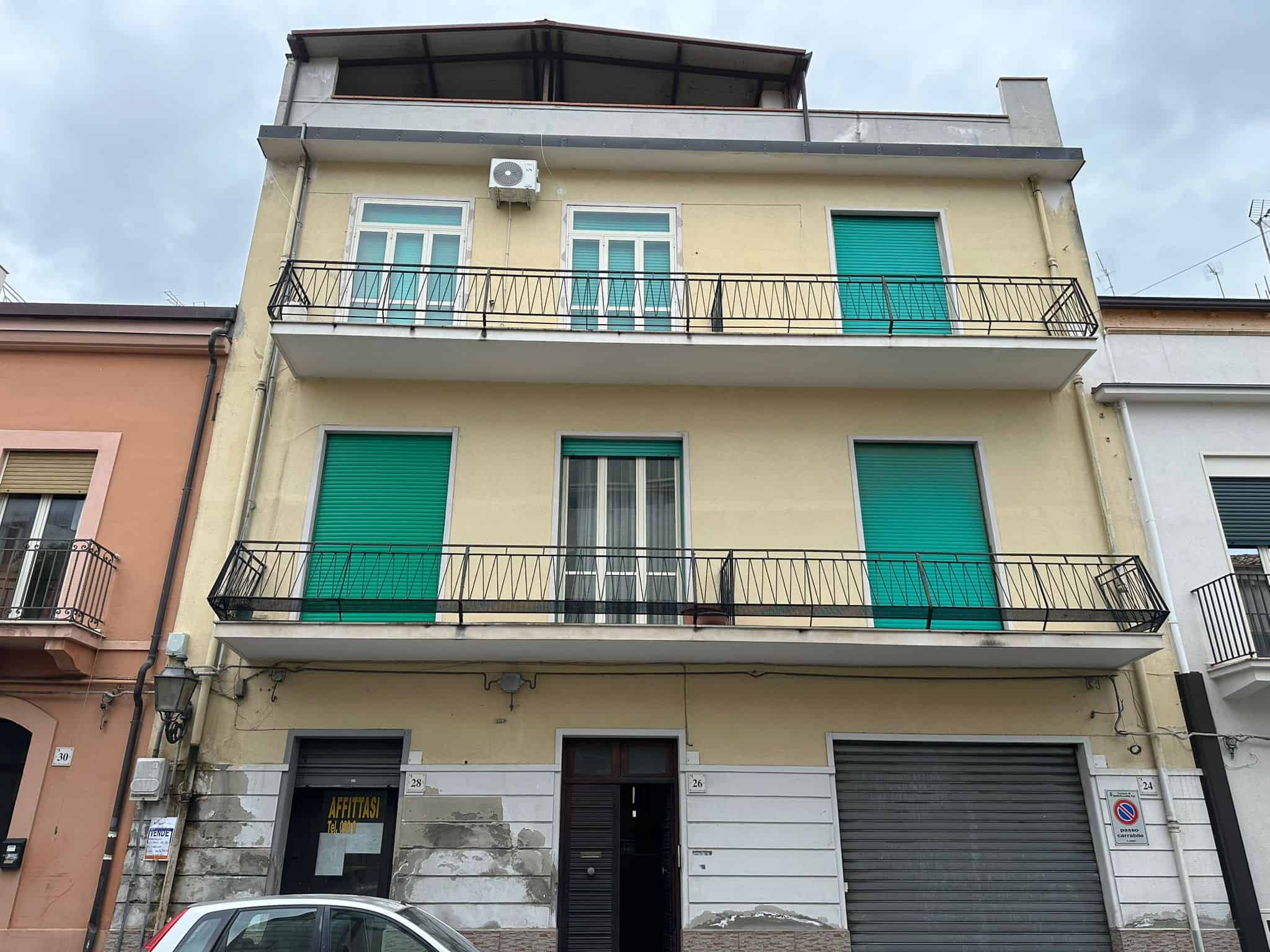 Balcone di un edificio residenziale con porte e finestre a vetri, persiane verdi, ringhiera in metallo e aria condizionata, situato in un'area urbana con altri edifici vicini.