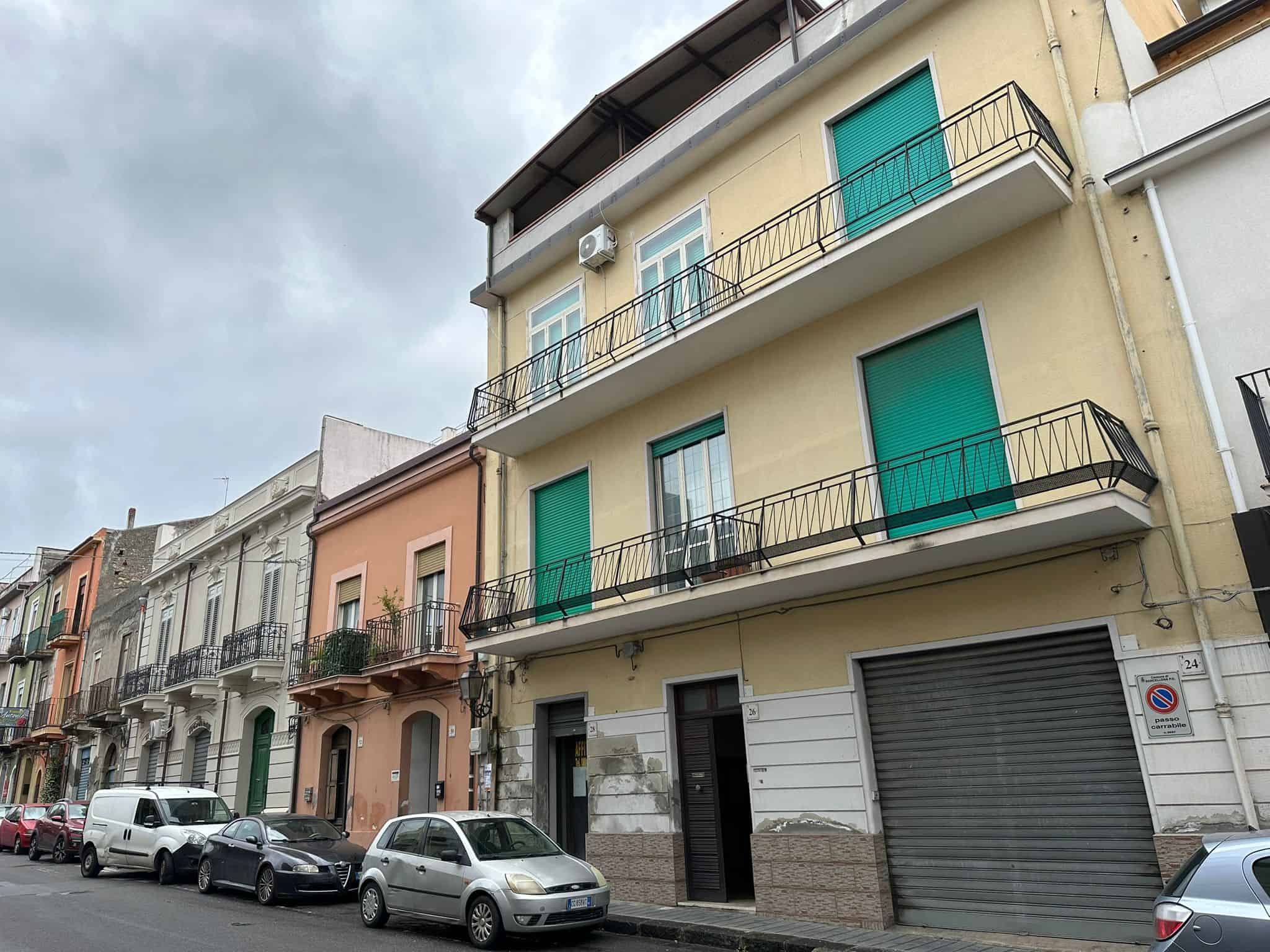 Finestra, balcone e facciata di un edificio residenziale in stile urbano con negozio al piano terra, su strada trafficata con auto parcheggiate, sotto un cielo nuvoloso.