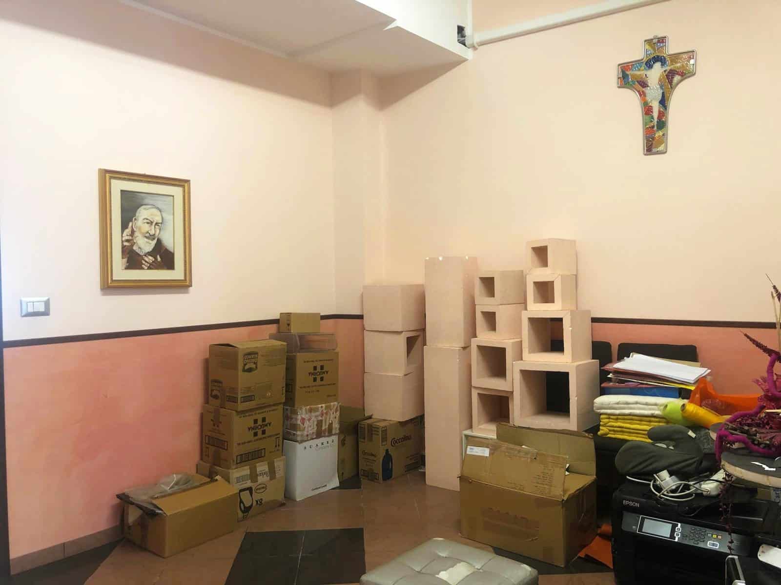 Scaffali in cartongesso e scatole di imballaggio in una stanza con pareti beige e rosa, decorata con un quadro e una croce colorata, pronta per trasloco o ristrutturazione.