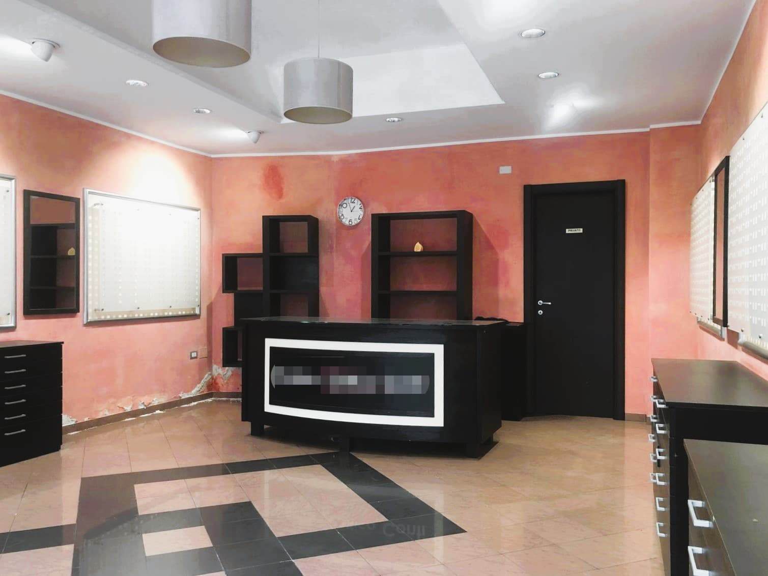 Receptione ufficio con pareti arancioni, arredi neri e scaffali aperti, illuminata da luci da soffitto e con un orologio a parete. Un ambiente moderno e funzionale, ideale per attività di accoglienza o lavoro.