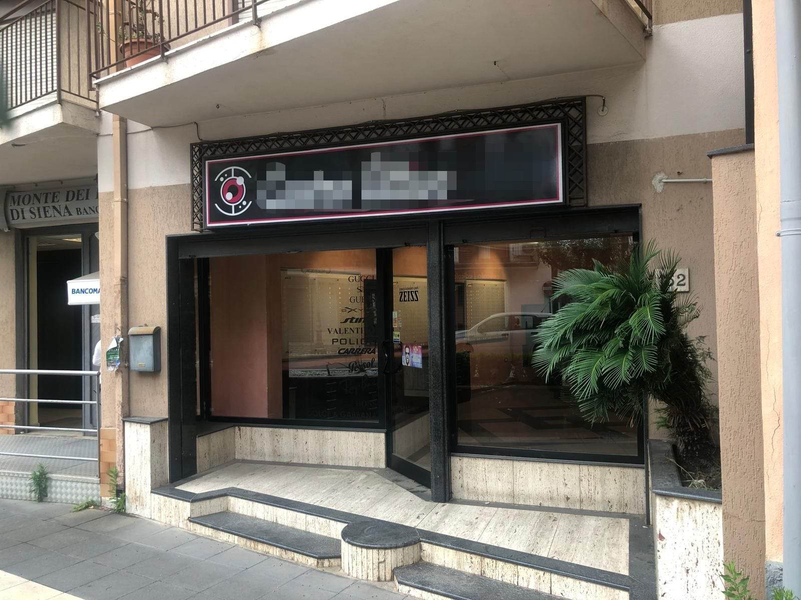 Vetrina commerciale con insegna digitale, ingresso con accesso pedonale in sala di forma rettangolare con grandi vetrate e una pianta ornamentale alla destra, in zona urbana.