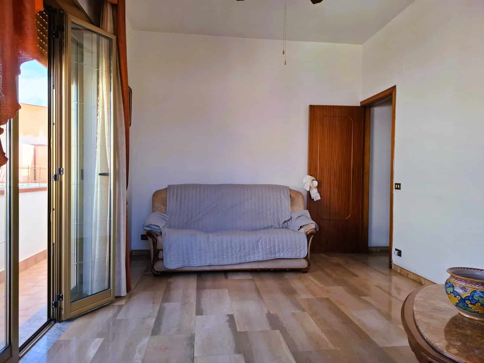 Spazio living luminoso con porte finestra che dà accesso al balcone, pavimenti in legno chiaro, divano in tessuto beige e dettagli in madera, ambiente accogliente e tranquillo.