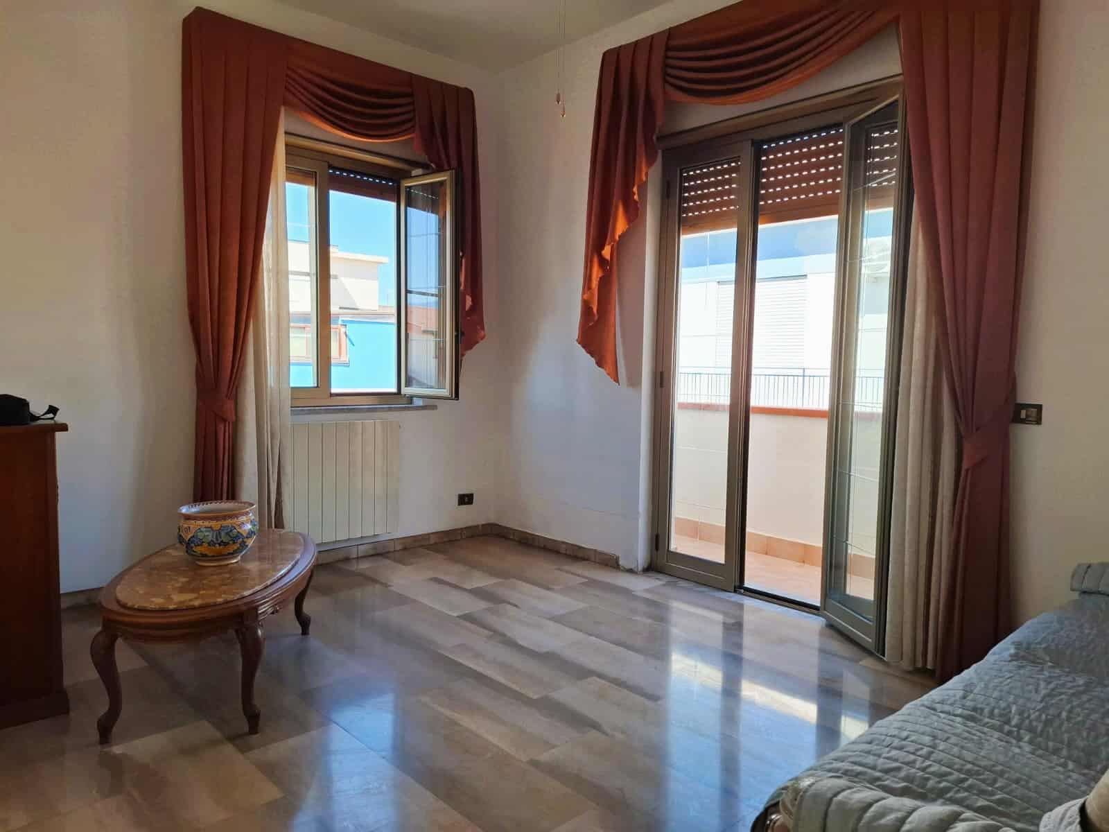 Soggiorno luminoso con grandi finestre e balcone, arredi classici, tende con doppio drappeggio color terracotta, pavimento in marmo beige, ideale per appartamenti di prestigio.