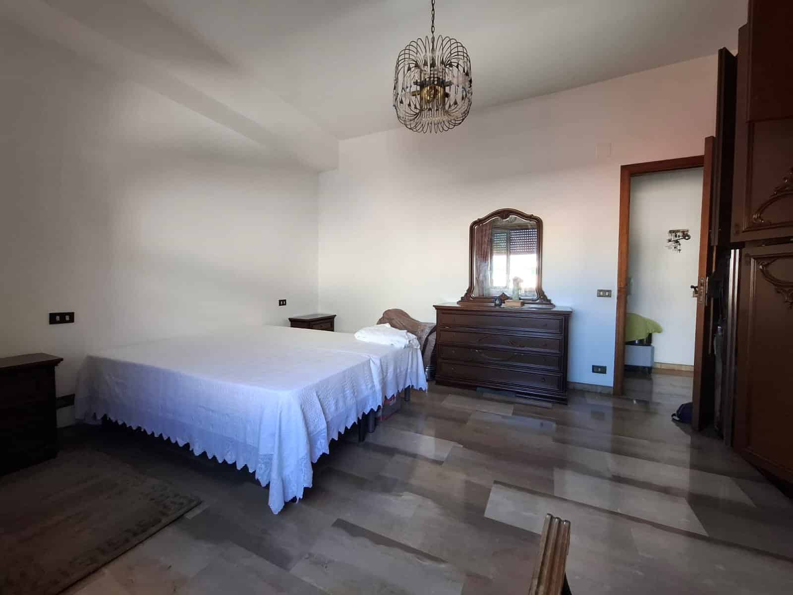 Letto matrimoniale in camera da letto con arredi in legno, lampadario elegante e pavimento in legno chiaro, ambiente spazioso e luminoso, ideale per vivere in comfort.