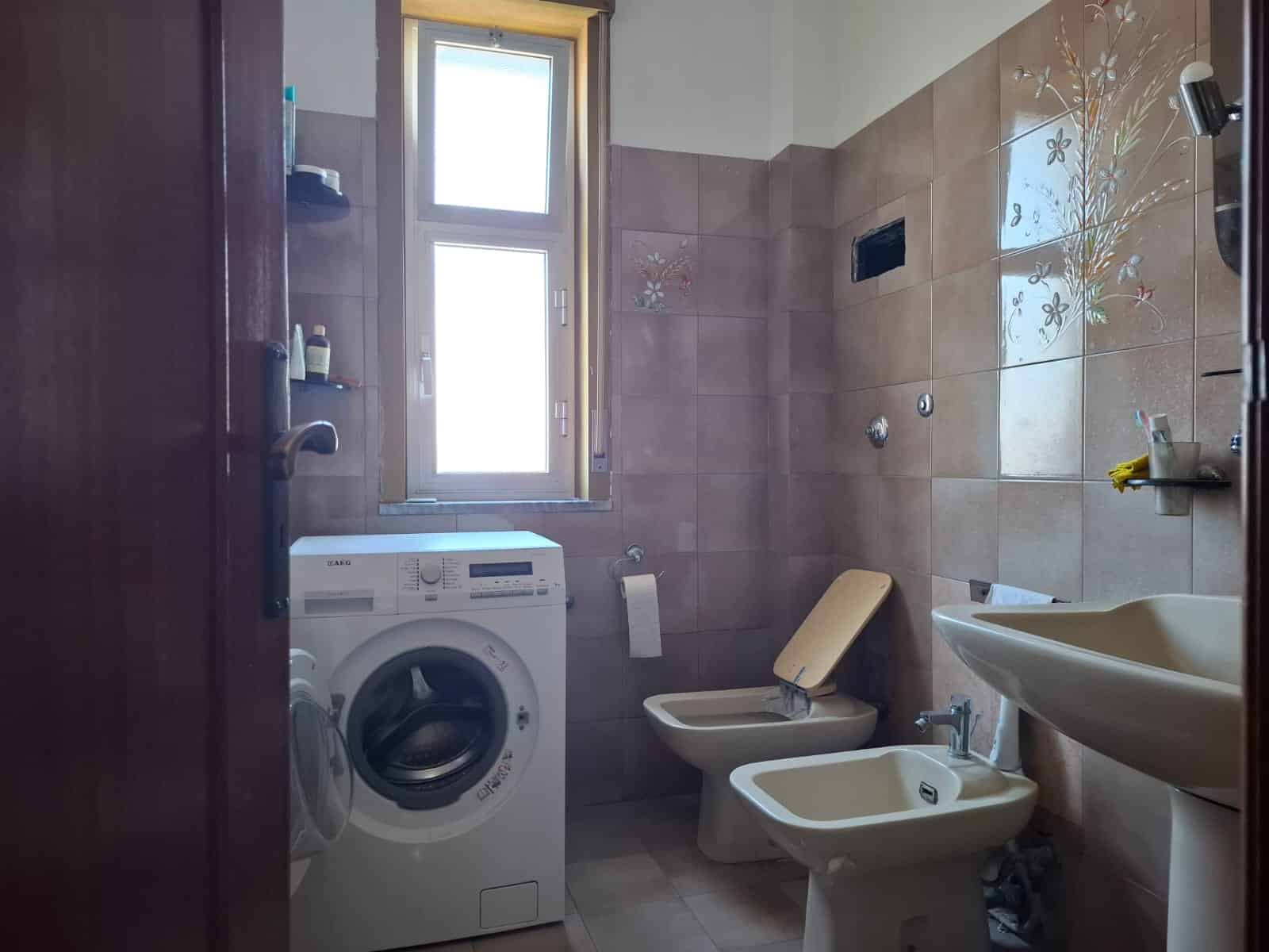 Lavanderia bagno con lavatrice, bidet, lavabo e finestra, arredato con piastrelle beige e decorazioni floreali, ideale per proprietà in vendita o affitto.
