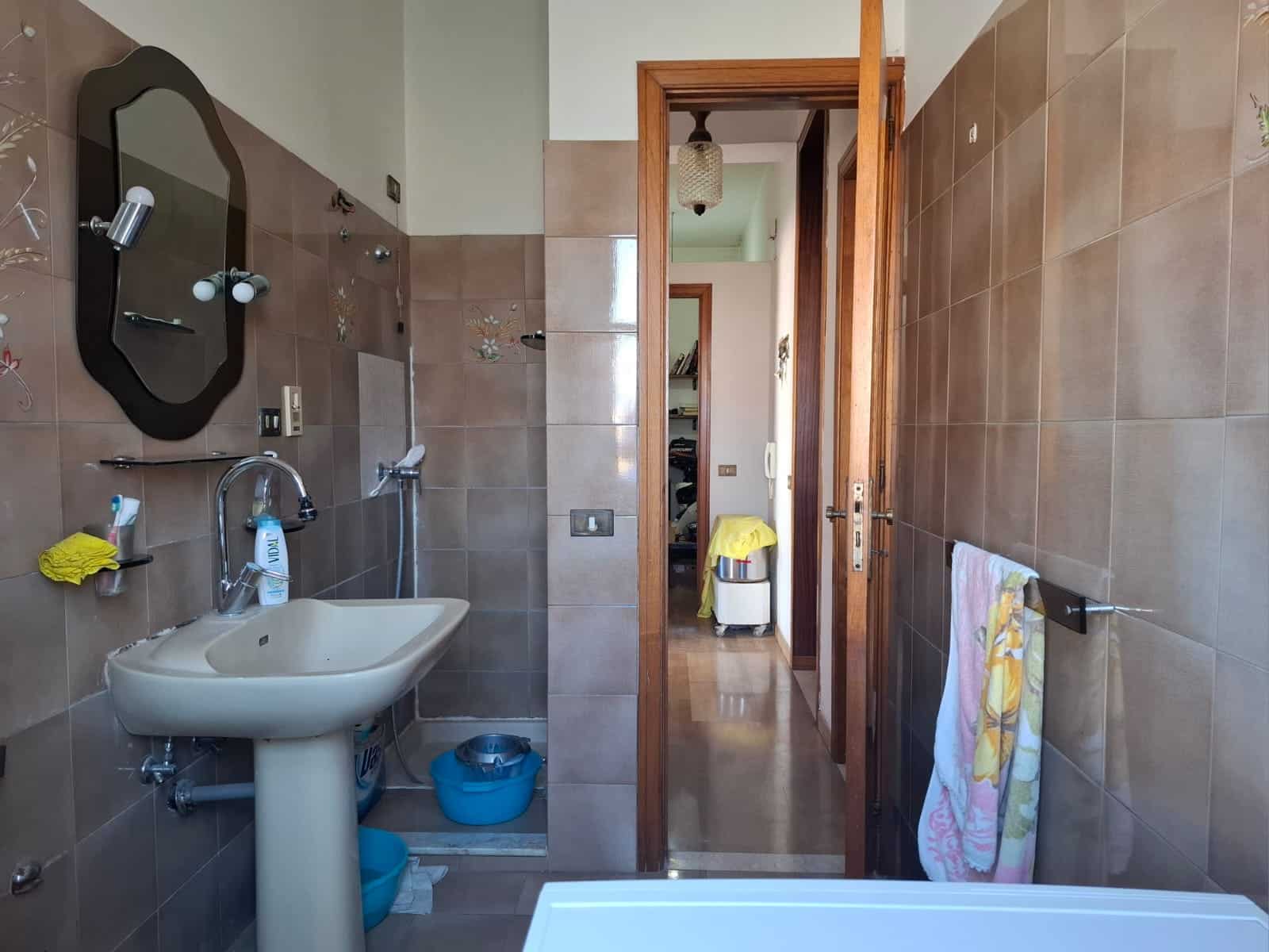 Lavabo e dettagli bagno con pareti rivestite in gres beige, specchio decorato, accessori bagno e asciugamani colorati. Vista verso la porta di accesso e un disimpegno con arredi.