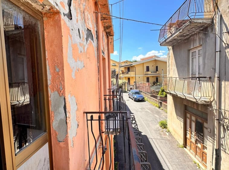 Balcone di un appartamento in condizione di deterioramento, con pareti dipinte di arancione e pareti che mostrano segni di peeling e muratura esposta, affacciato su una strada urbana soleggiata.