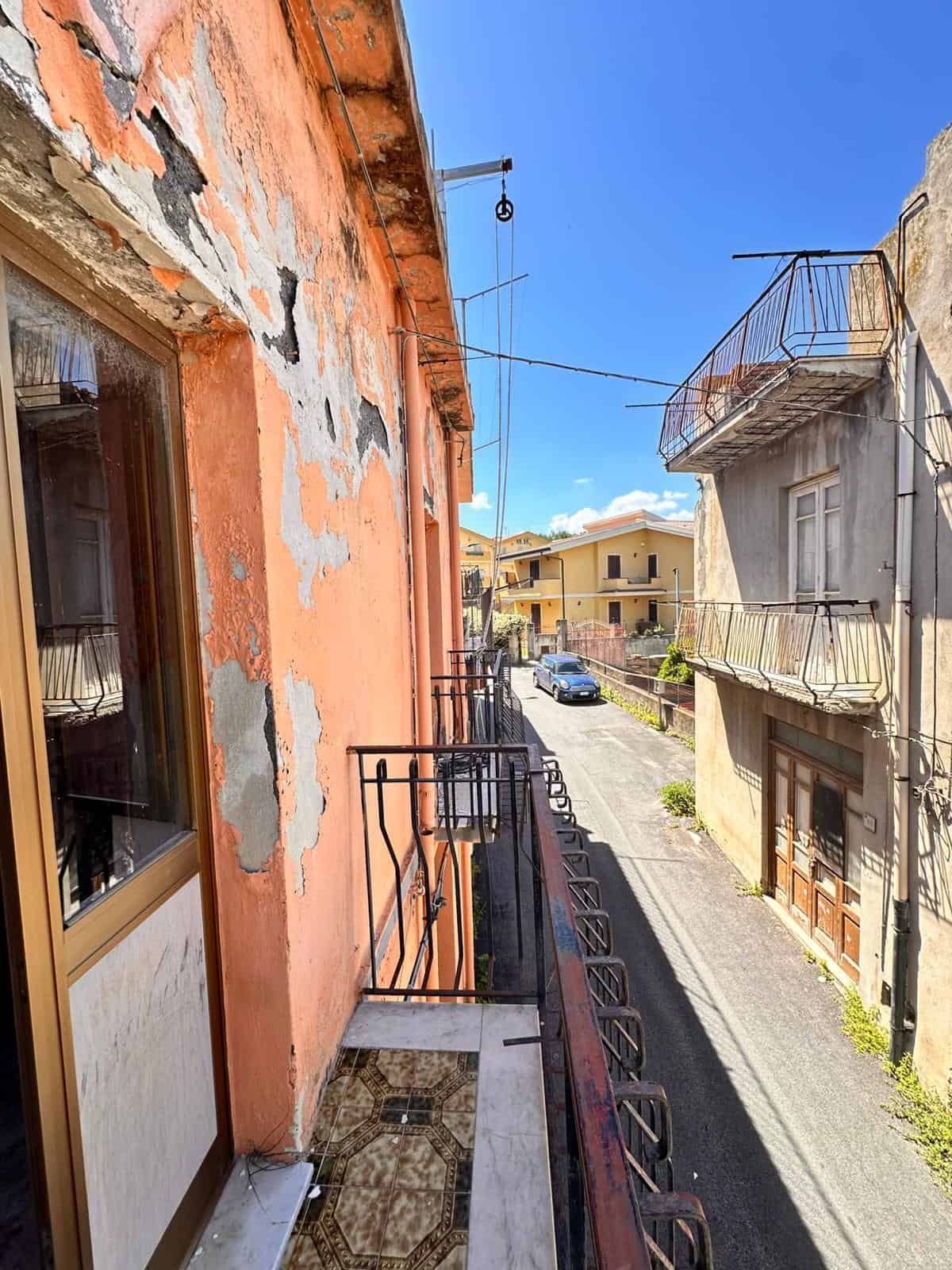 Balcone di un appartamento in condizione di deterioramento, con pareti dipinte di arancione e pareti che mostrano segni di peeling e muratura esposta, affacciato su una strada urbana soleggiata.