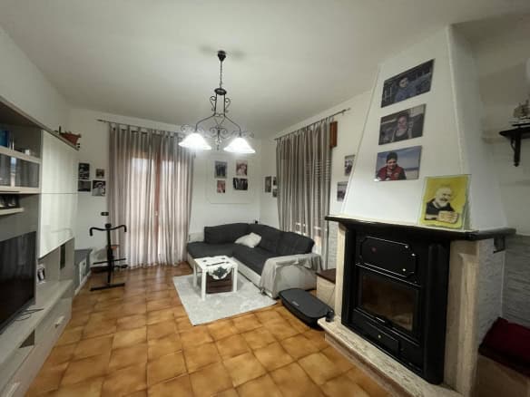Ampio soggiorno con camino, divano nero e pareti decorate con foto di famiglia, illuminato da lampadario centrale e grandi tende trasparenti, ideale per vivere momenti di relax e convivialità.