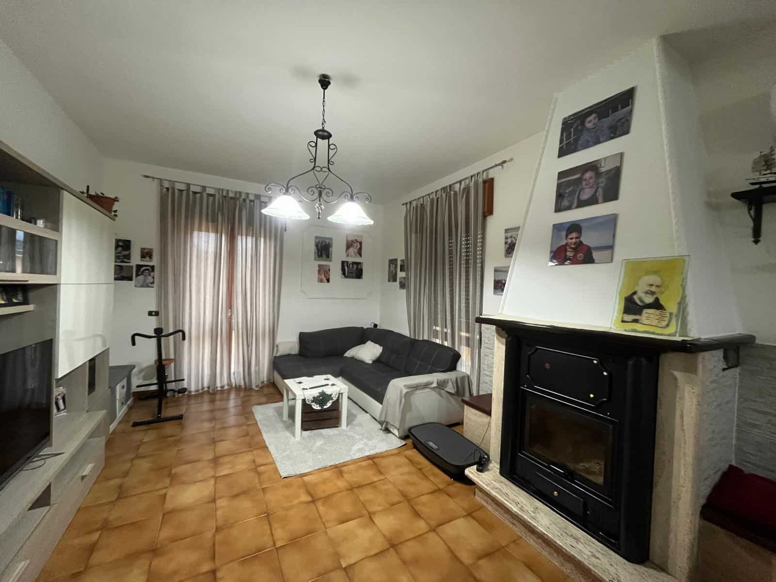 Ampio soggiorno con camino, divano nero e pareti decorate con foto di famiglia, illuminato da lampadario centrale e grandi tende trasparenti, ideale per vivere momenti di relax e convivialità.