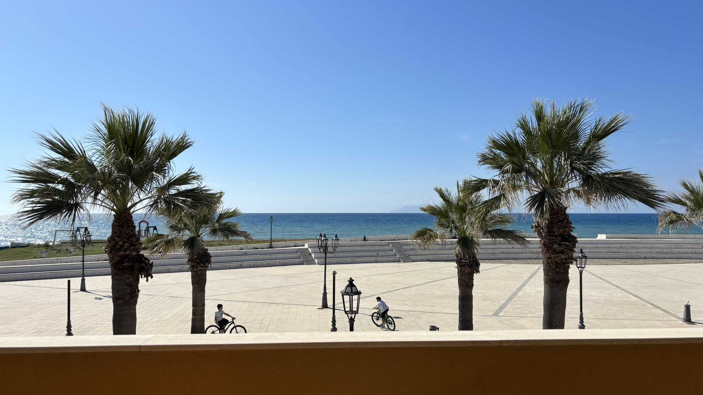 Vighevoli di palme vista mare sulla passeggiata lungomare con ciclisti, lampioni e cielo sereno, ambiente Mediterraneo, immagine di alta qualità per promuovere case vacanza e proprietà immobiliari sul mare.