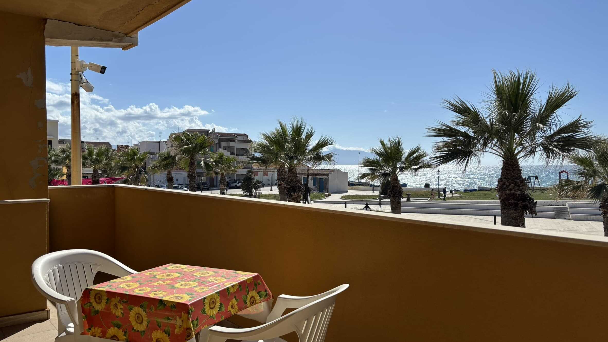 Balcone con vista mare, ombra e tranquillità, arredato con tavolo e sedie in plastica, palme e mare sullo sfondo, ideale per relax e svago in estate, casa al mare NextCasa.