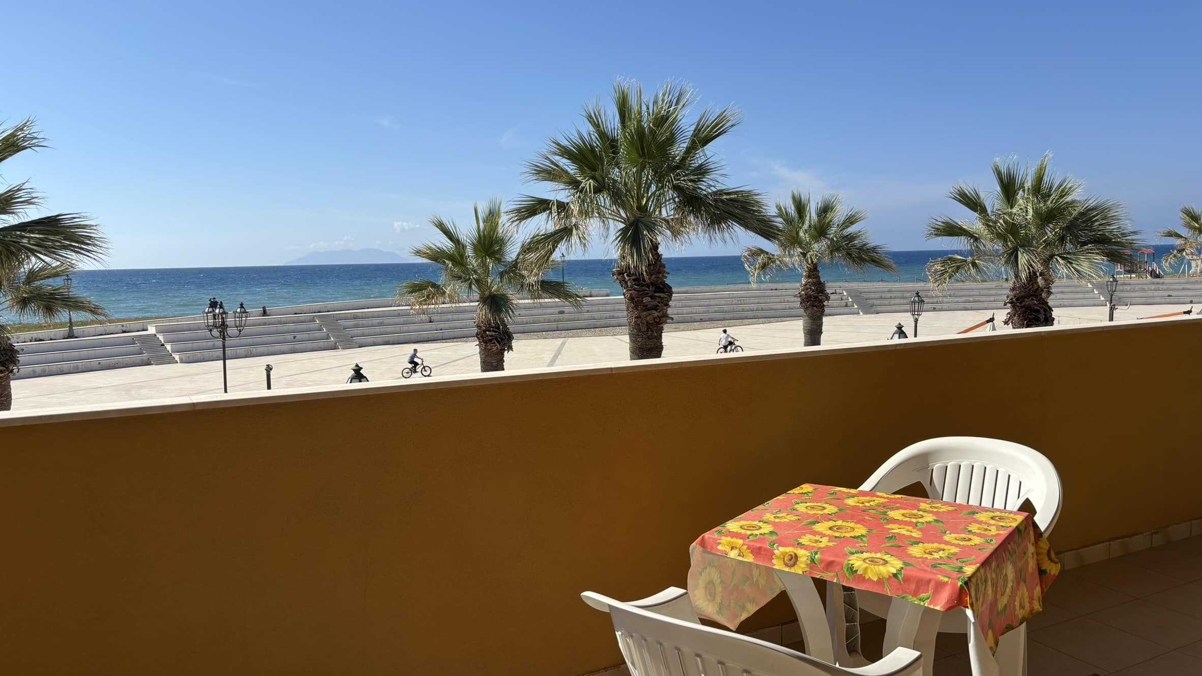 Balcone vista mare con tavolo con tovaglia a fiori, sedie di plastica, palme, spiaggia e cielo blu in background. Perfetto per vacanze al mare e relax.