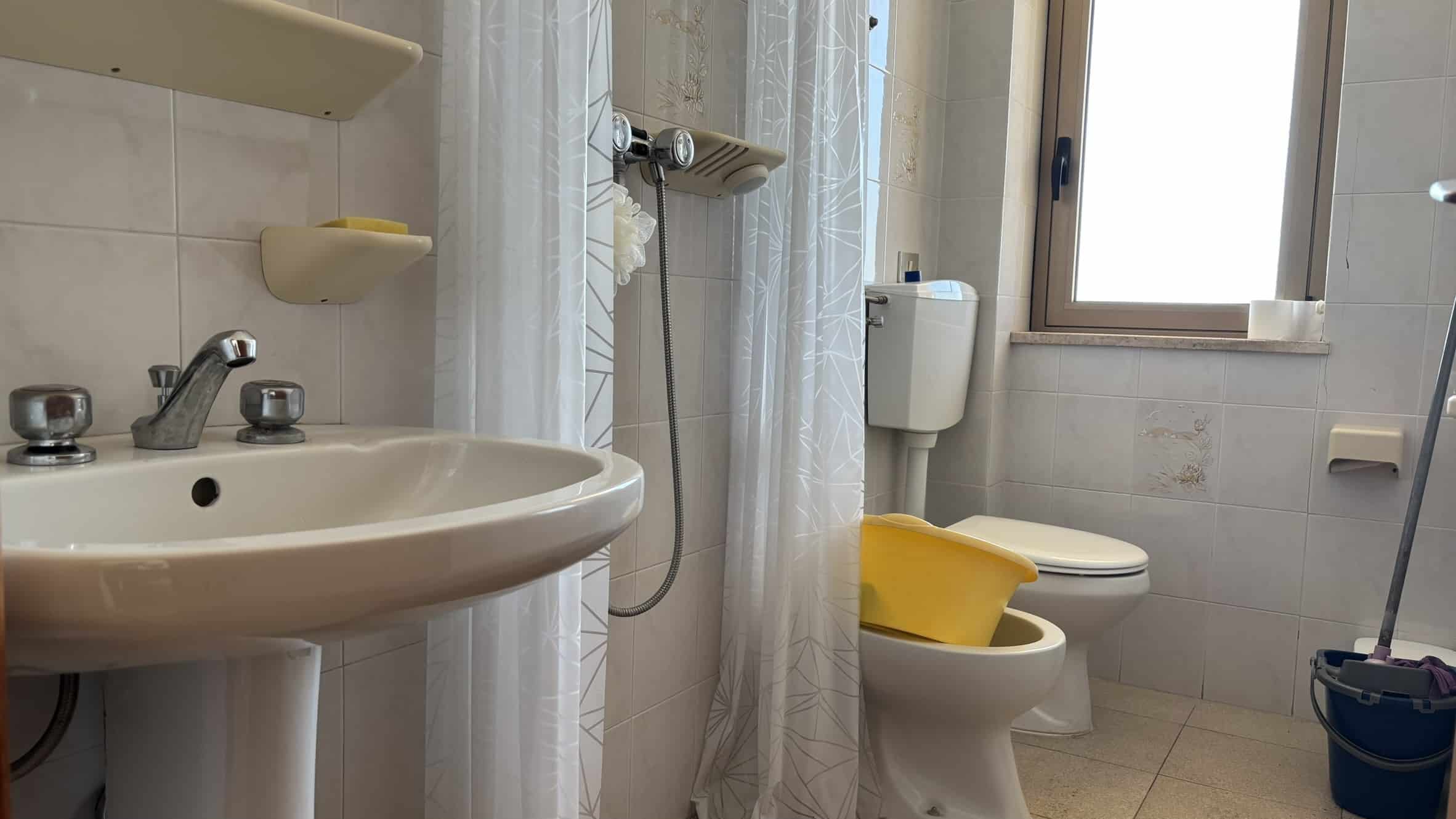 Lavabo bagno con angolo doccia, finestra e servizi igienici, arredi semplici e funzionali, ideale per ambienti di abitazione, alloggi di NextCasa in Italia, bagno pratico e luminoso.