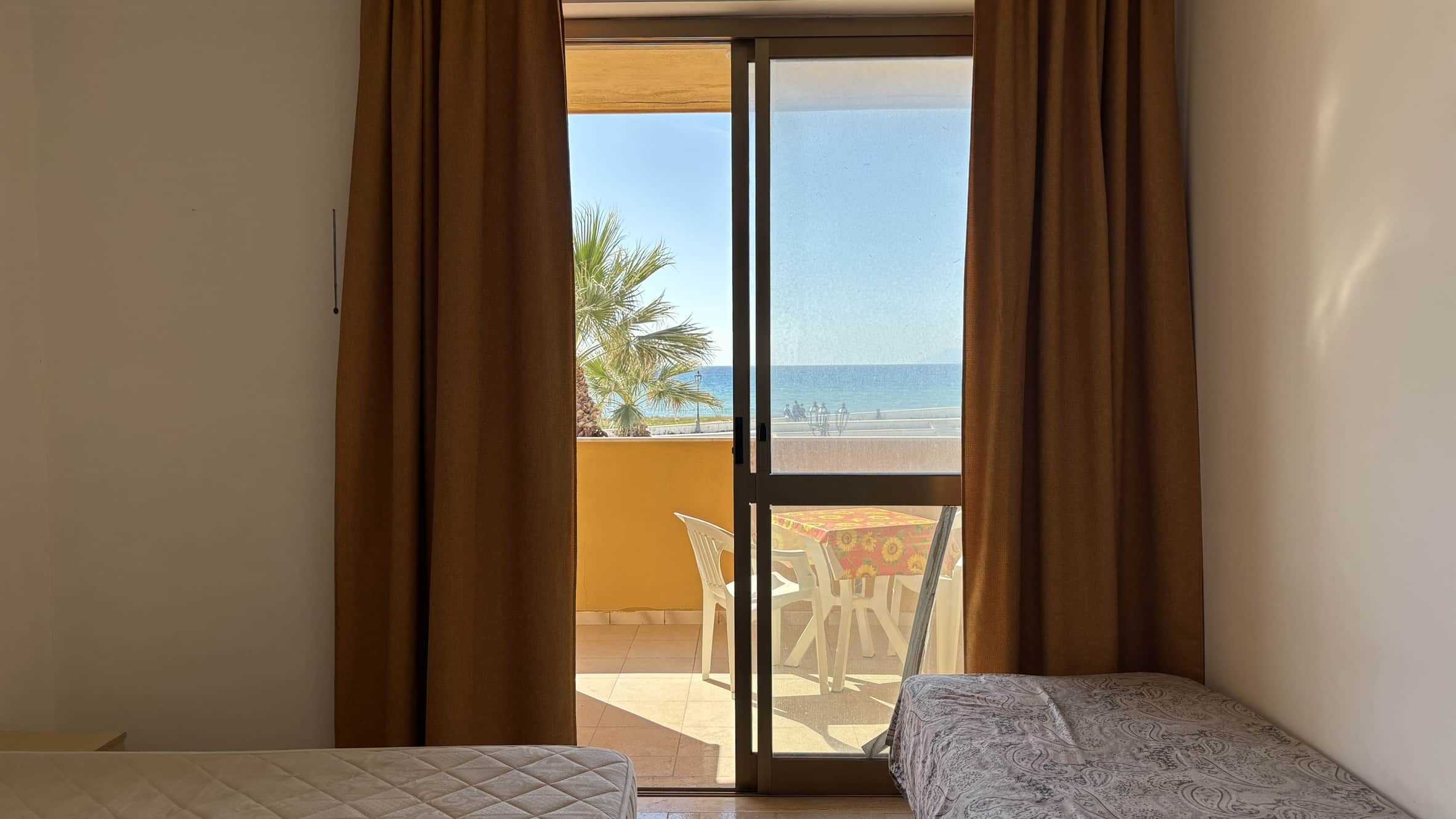 Vista mare dalla camera con balcone, arredamento semplice, tende marroni, pensata per case vacanza vicino al mare, con accesso diretto alla spiaggia, ideale per soggiorni rilassanti e confortevoli.