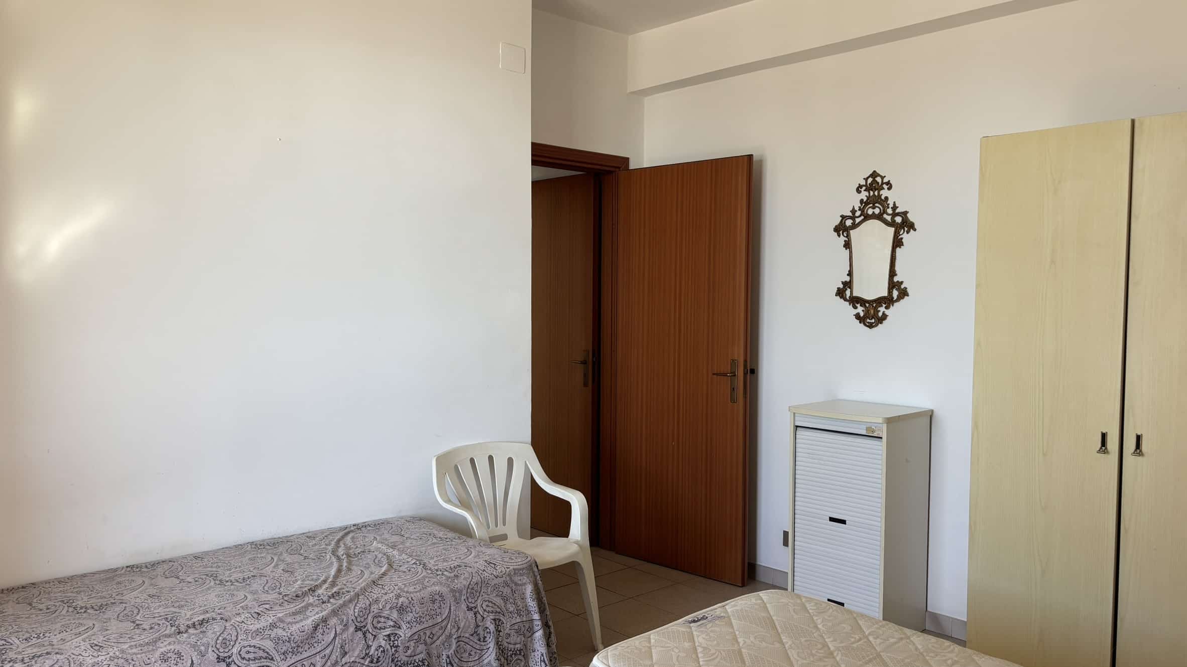 Letto singolo con copriletto in tessuto decorativo, armadio e specchio antico, ambiente camera semplice e funzionale, ideale per affitto residenziale o studenti.
