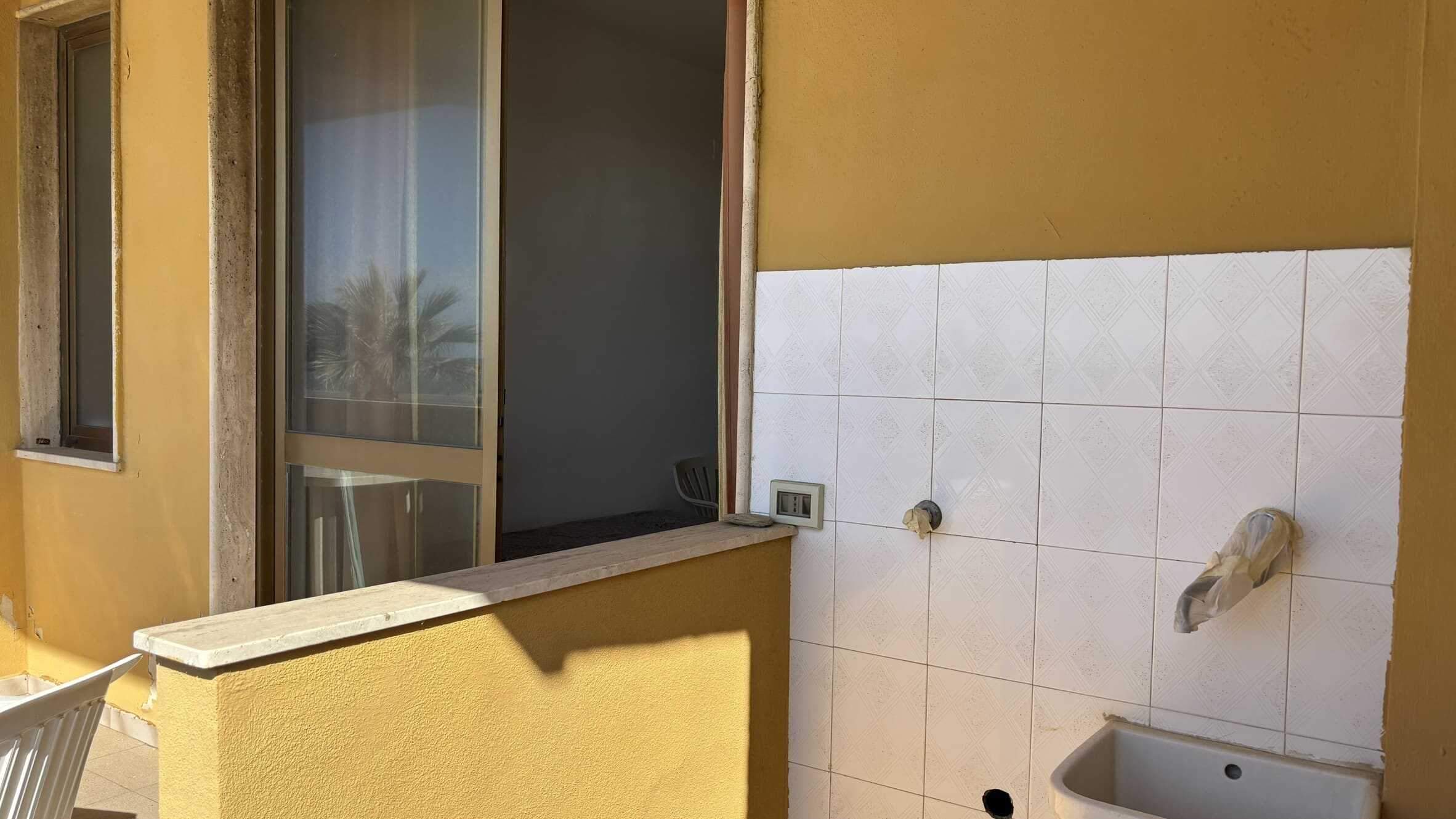 Rifacimento balcone con vista su palma, pareti gialle e mattonelle bianche, pronto per installazione lavandino e rubinetto, ambienti luminosi e ventilati, ideale per rinnovare la zona lavanderia.