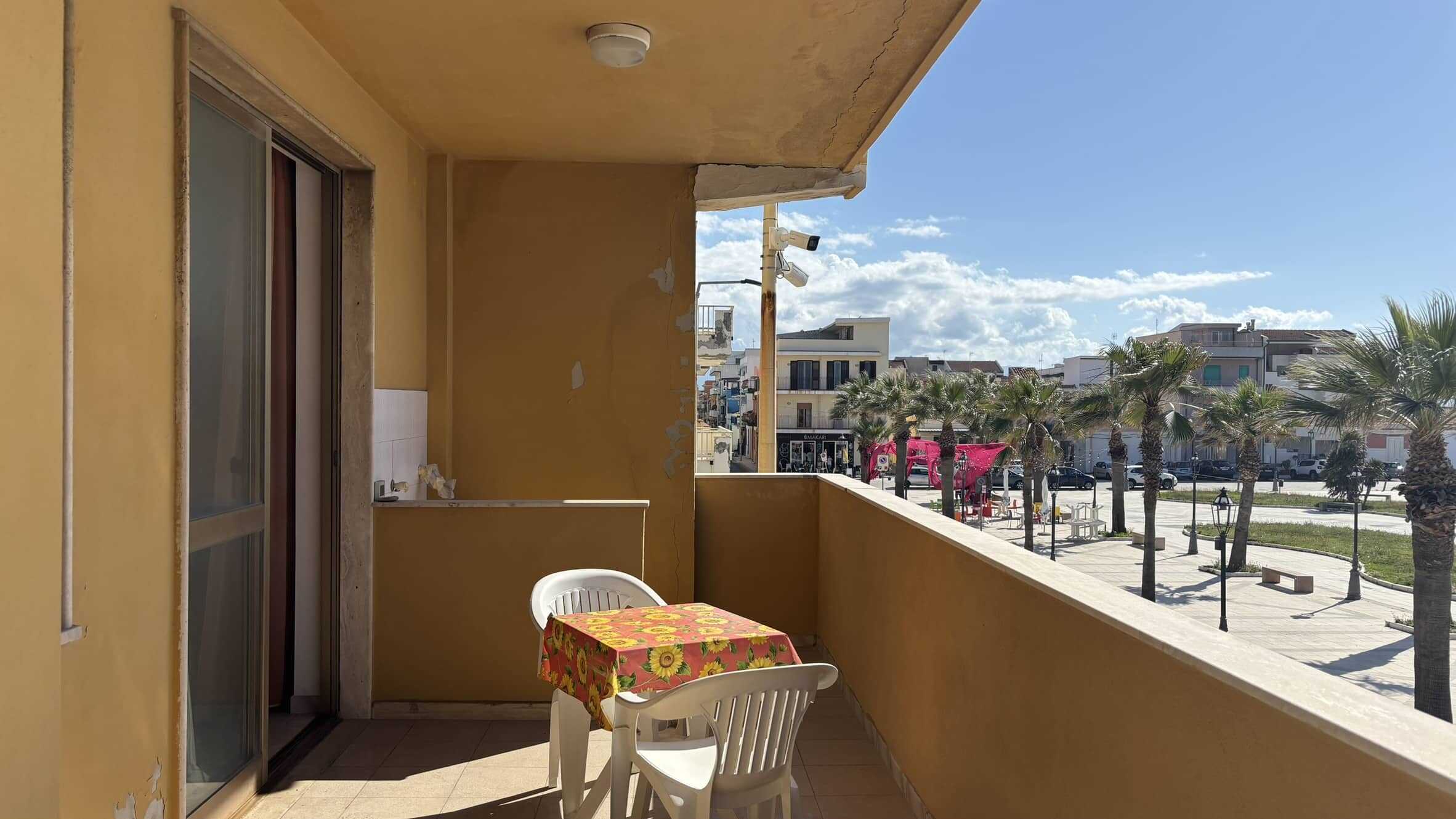 Balcone di un appartamento a mare con vista sulla spiaggia e palme, arredato con tavolo e sedie, edificio color giallo, cielo blu e clima soleggiato, ideale per vacanze al mare.